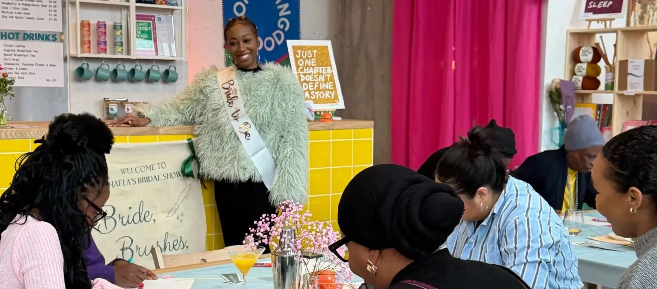 A woman wearing a sash and a fluffy sweater standing behind a decorated table at a bridal shower, surrounded by guests sitting and painting at the table.