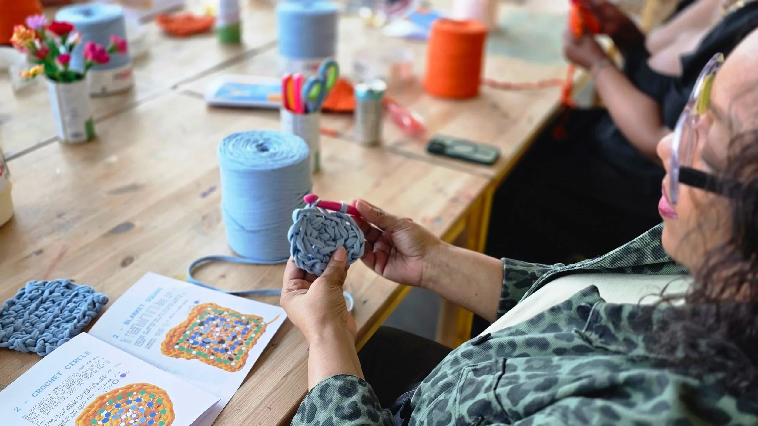 A woman in a camouflage jacket and safety glasses learning to crochet at a wooden table. The table has colorful yarn, a craft manual, and finished or in-progress crochet projects. Several cans of colorful thread or yarn are arranged on the table.