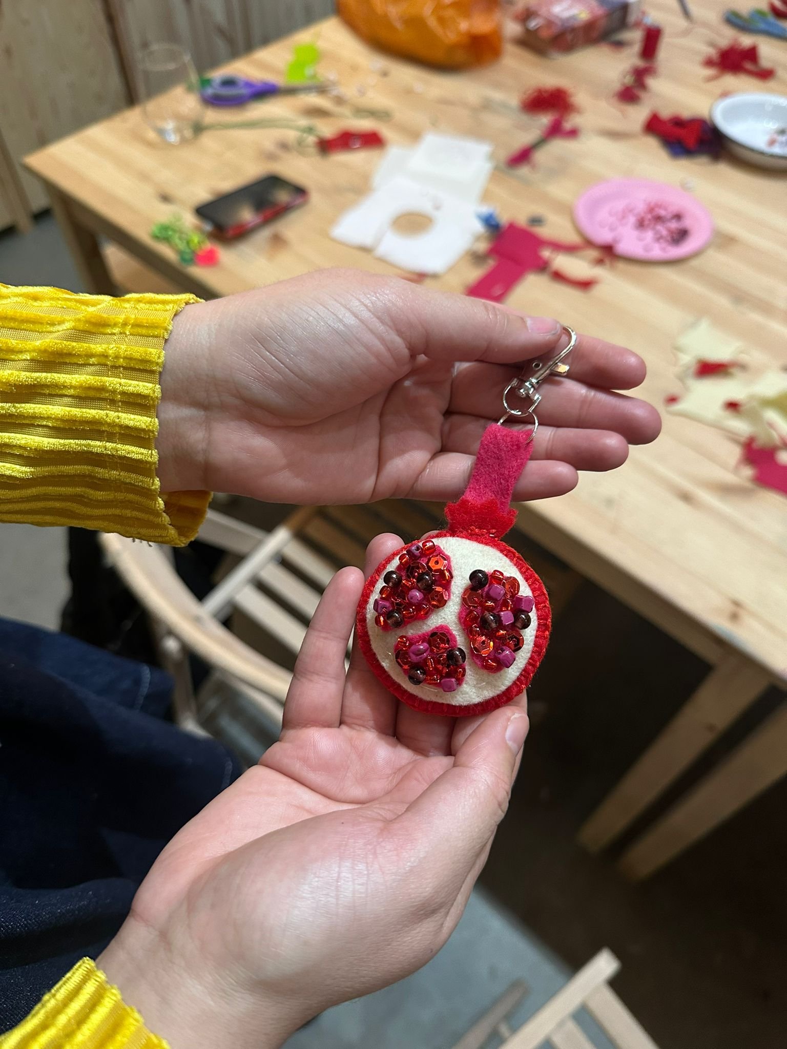 A person's hand holding a felt ornament decorated with small beads and sequins, with a craft workshop in the background.