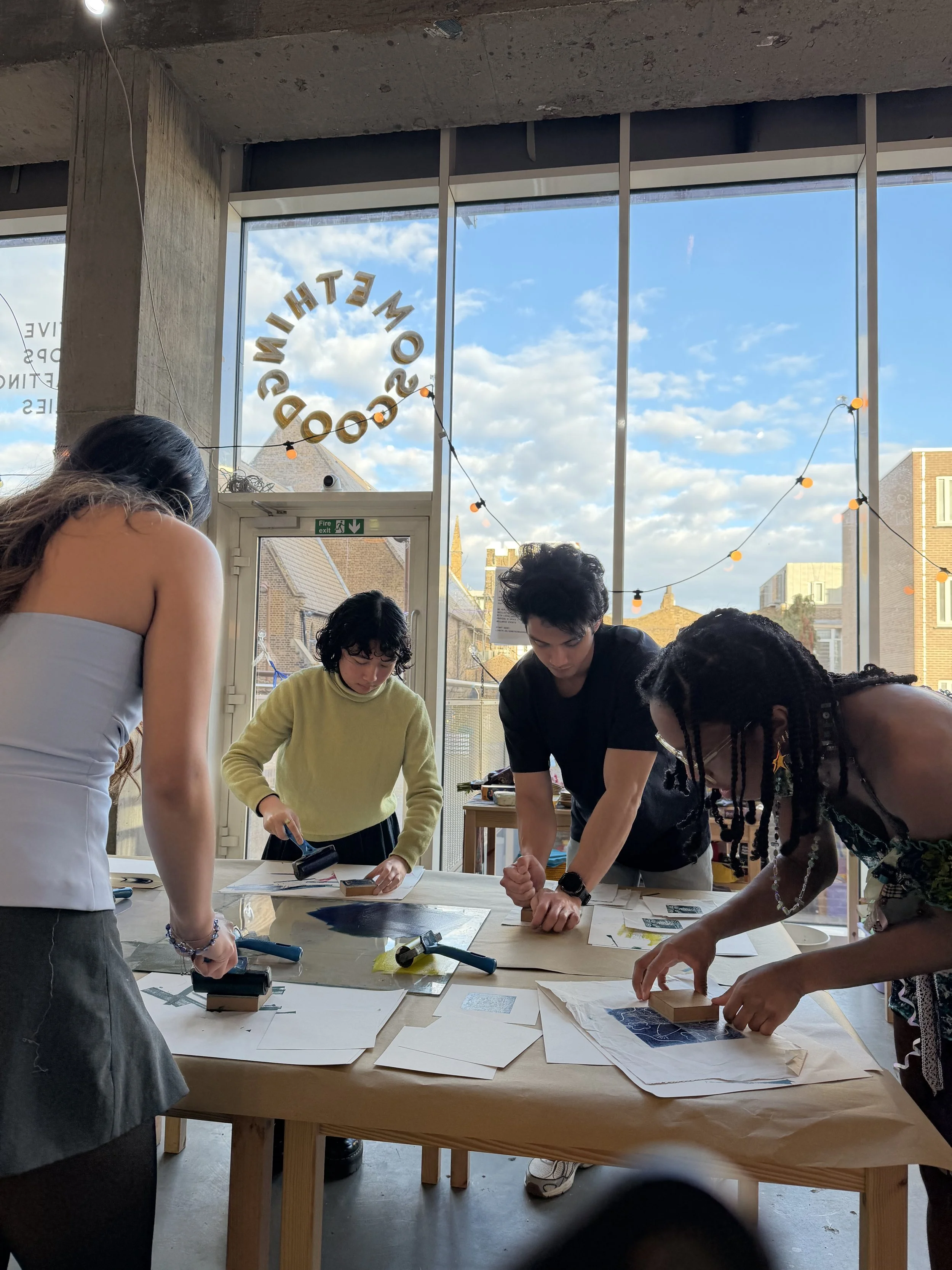 A group of people standing around a large table covered with papers and tools, working on a project inside a room with large windows showing a blue sky with clouds outside at Something Good Workshops in Deptford, London. 
