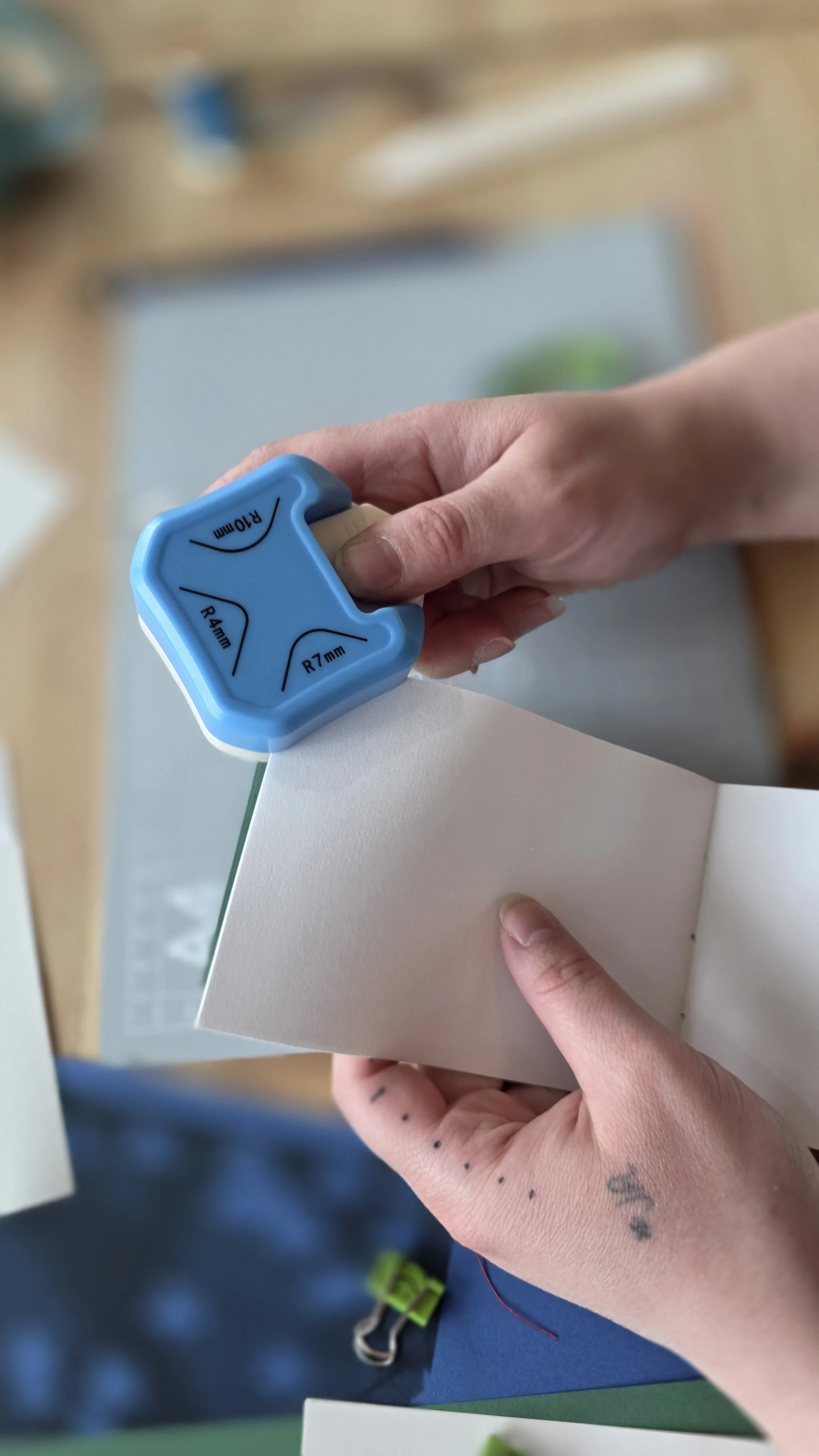 Person pressing a corner rounder tool against the corner of a white sheet of paper at a bookbinding workshop in London.