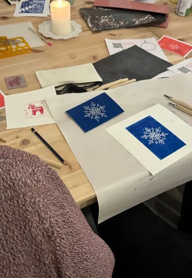 Workspace with holiday card preparations, featuring blue lino slabs with snowflake designs, pens, markers, and a lit candle.