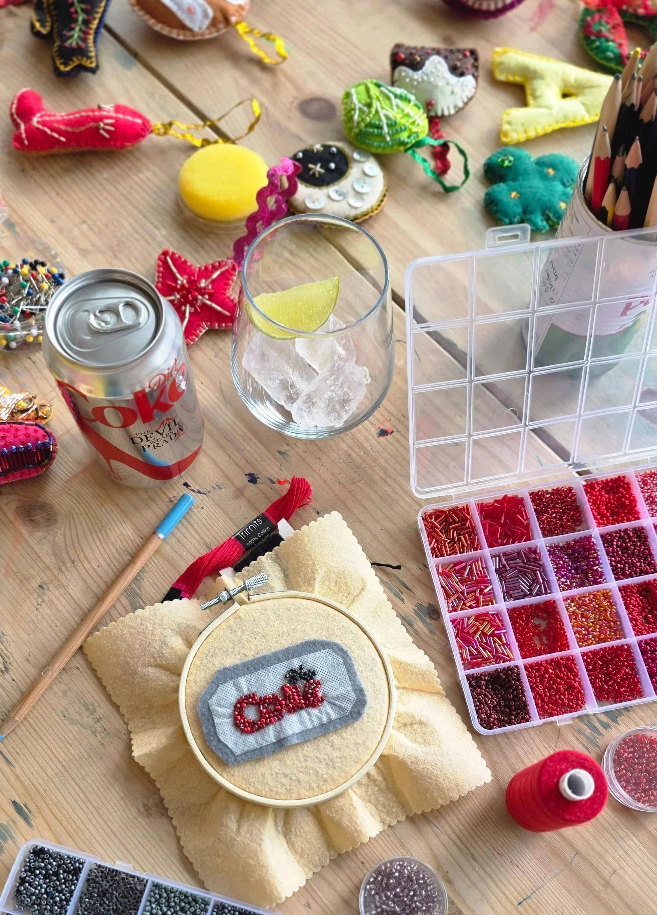 A craft table with various colorful felt ornaments, a container of small colored beads, a can of Diet Dr. Pepper, a glass of ice with a lemon wedge, and a cross-stitch project spelling 'Candy.'