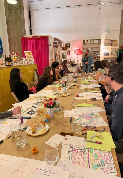 People attending a workshop at a long table filled with papers, wine glasses, drawings, and art supplies in a cozy indoor setting.