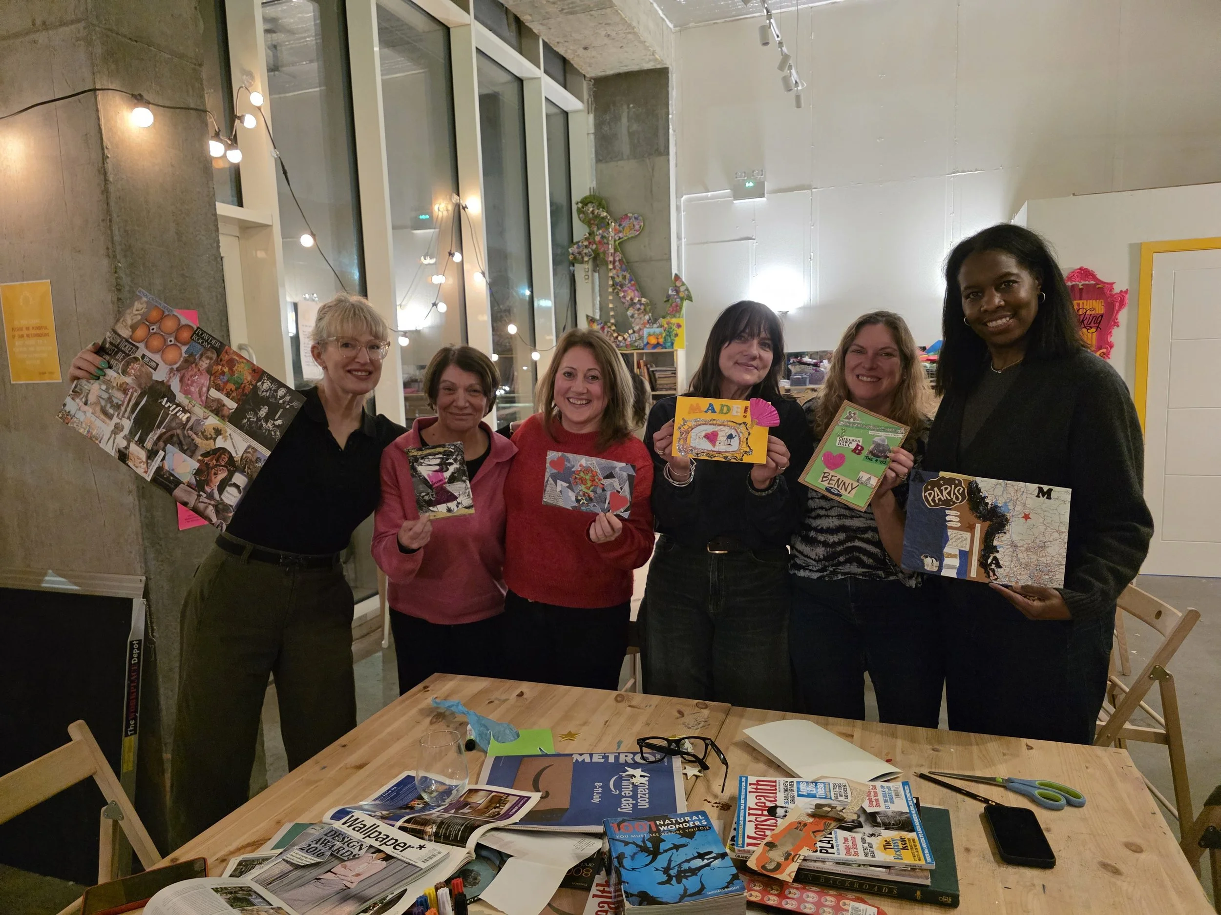 Six women are standing and smiling for a photo in a room with large windows, string lights, and creative artwork. They are holding small, decorated cards or pieces of artwork. The table in front of them is scattered with magazines, papers, scissors, and other craft supplies.