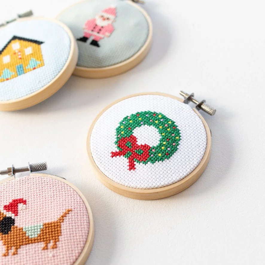 Cross-stitch embroidery hoops with holiday-themed designs, including a Christmas wreath with a red bow, a Santa Claus, a dog with a red Santa hat, and a house with snow.