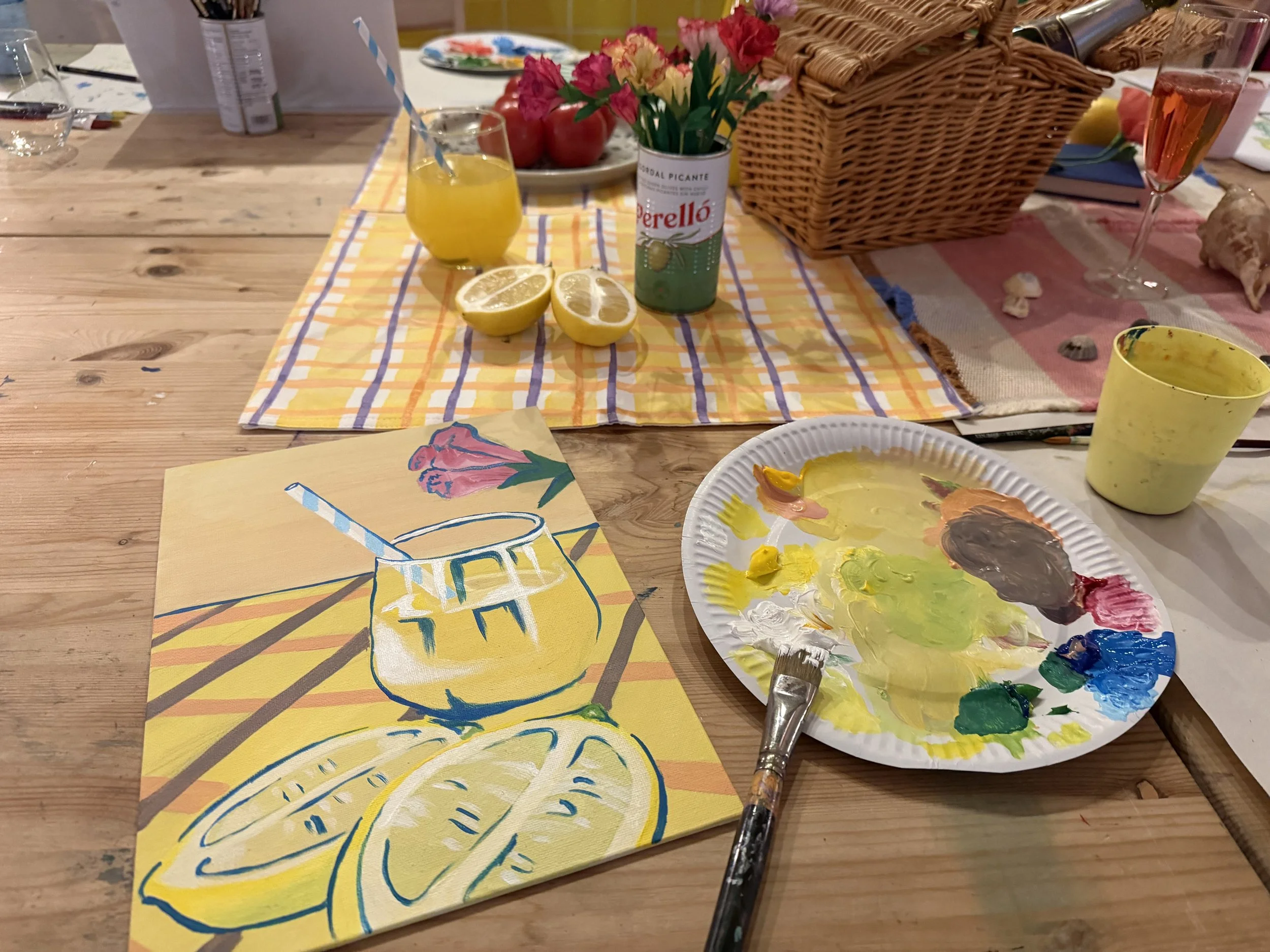 A wooden table with art supplies, including a painted sheet of paper with a yellow lemon slice, a paintbrush, a paper plate with yellow, green, and brown paint, a yellow cup, and a paper napkin featuring a pink flower design. In the background, there