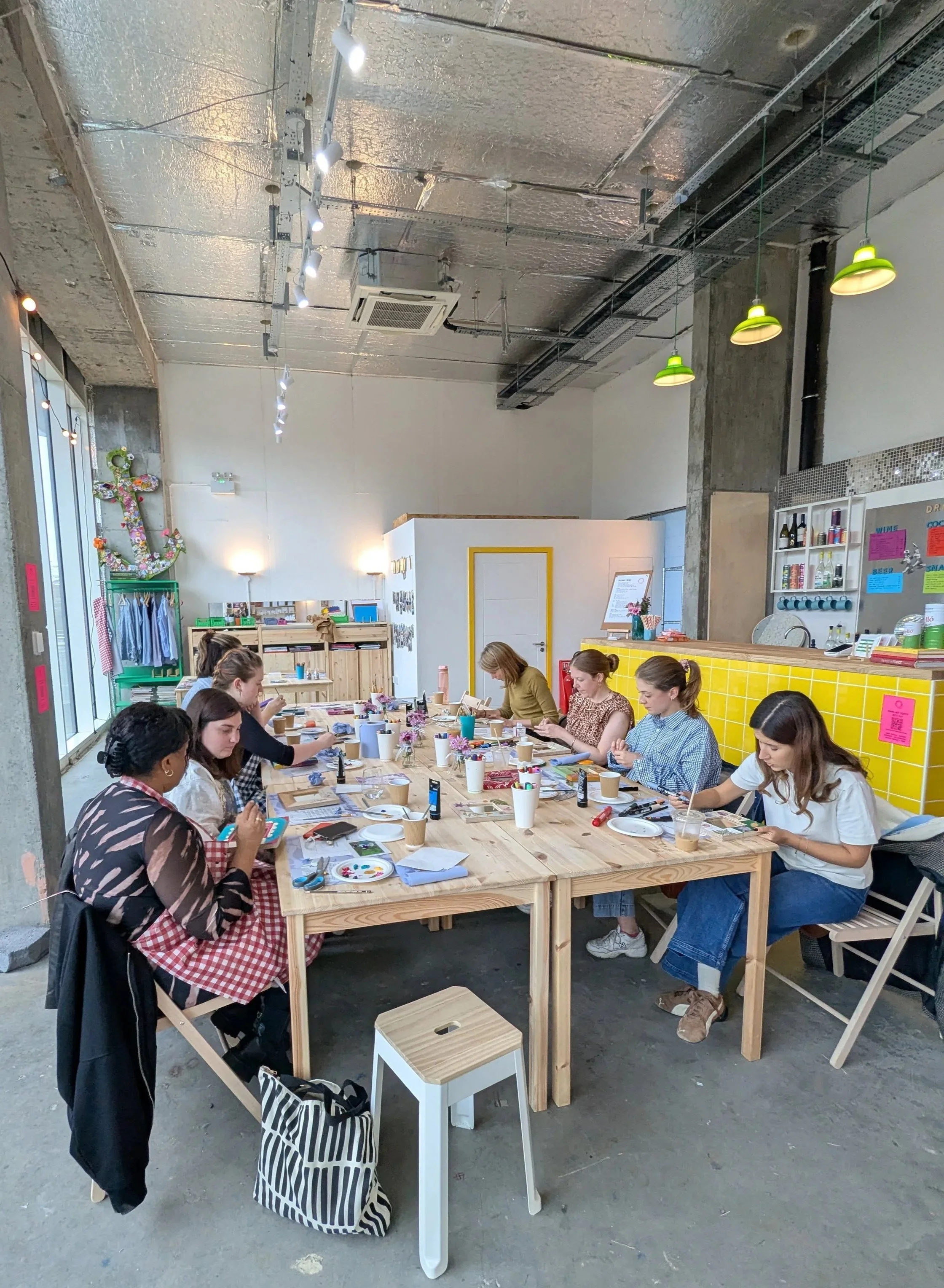 Inside a modern cafe or workshop, several women sit at a large wooden table engaging in various activities like drawing or organizing materials, with art supplies and cups in front of them. The ceiling has exposed metallic and industrial elements, with pendant and track lighting. The space includes a yellow-tiled counter, shelves with bottles, and a decorative anchor with flowers on the wall.