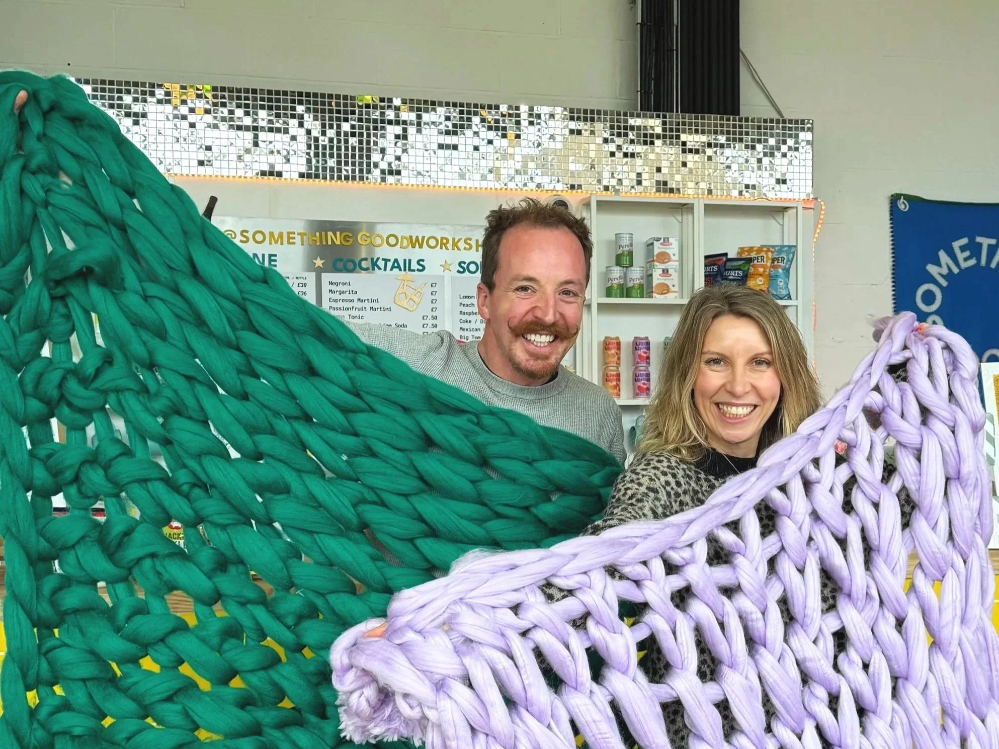 Two smiling people, a man and a woman, stand behind large, colorful, arm knitted blankets in a bright indoor space at Something Good Workshops in Deptford.