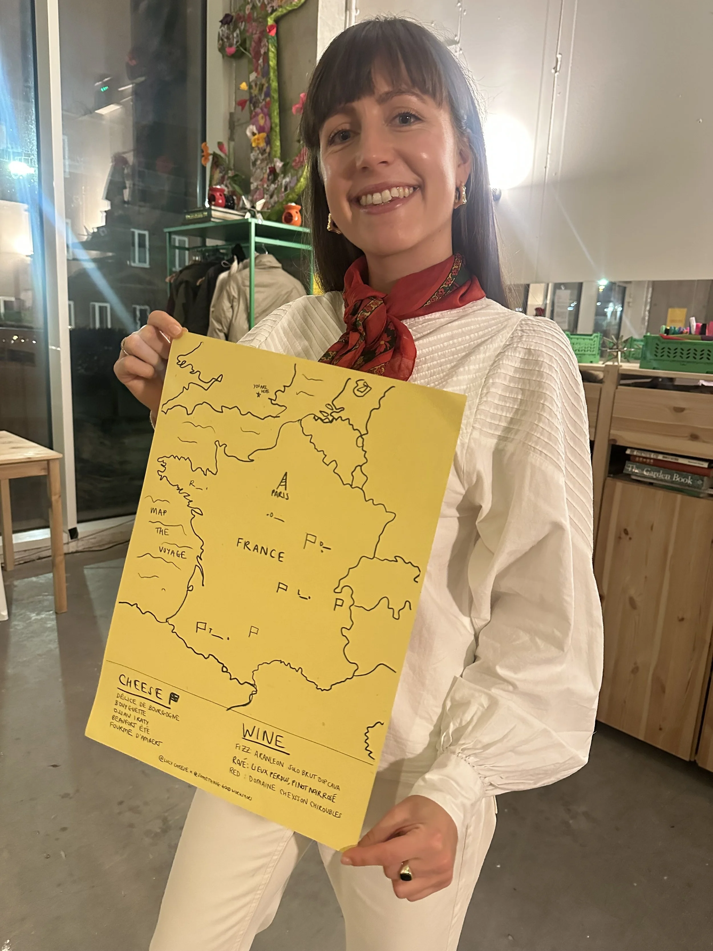 A woman smiling and holding a yellow map of France with handwritten notes on various regions, indoors at a cozy space with shelves and decorations at Cheese tasting event in London.