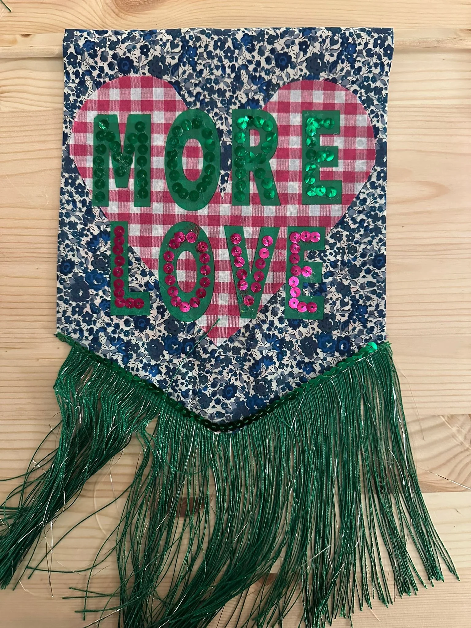 Decorative fabric banner with the phrase "Love More" written on it. The banner has a floral pattern background with a heart shape behind the text. The words are made of shiny green and pink sequins, and green fringes hang from the bottom edge.