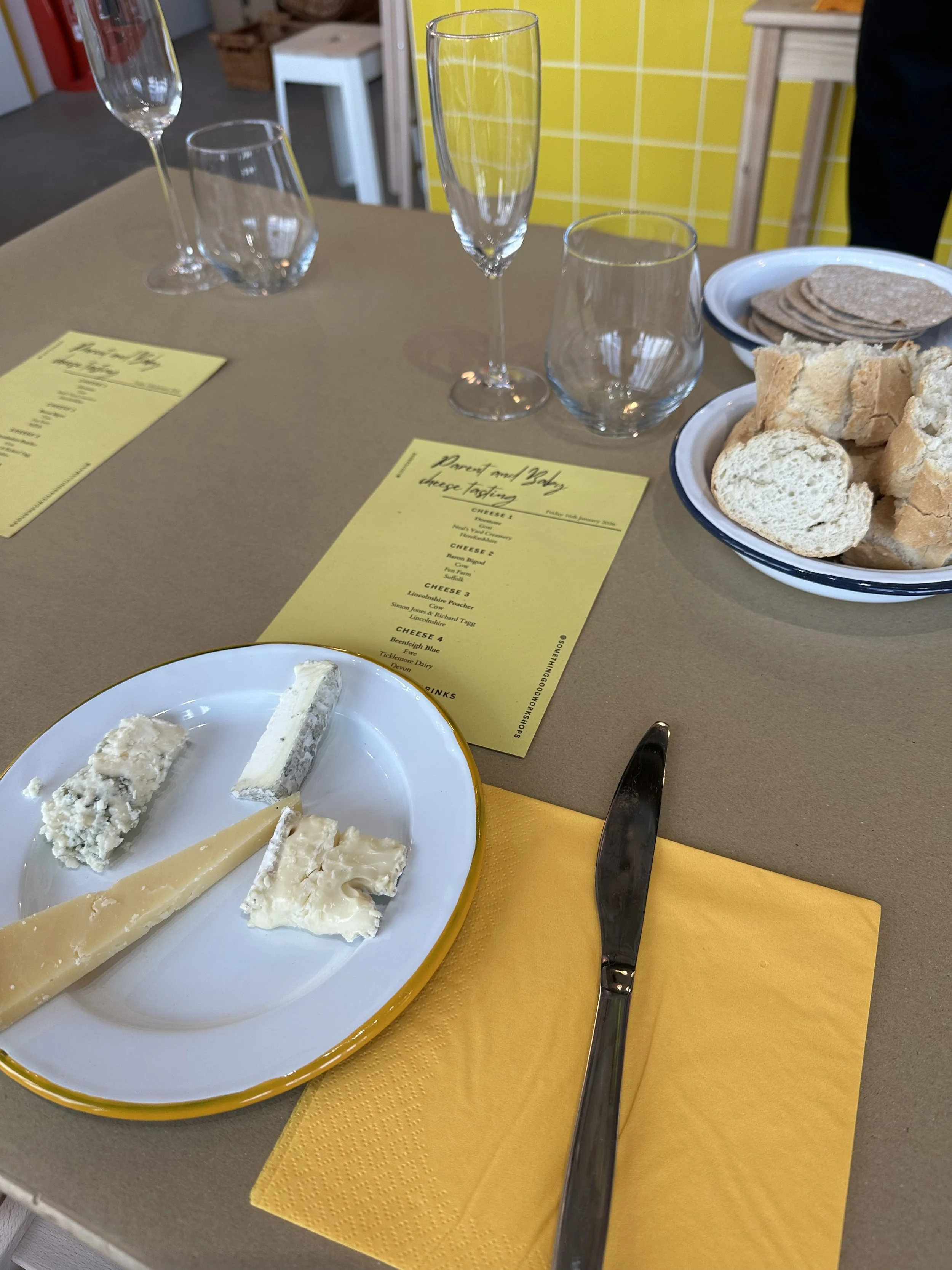 A cheese tasting arrangement with various cheeses on a white and yellow plate, a cheese tasting menu, dessert with a slice of cheesecake, wine glasses, a bowl of bread and crackers, placed on a table with a yellow napkin.
