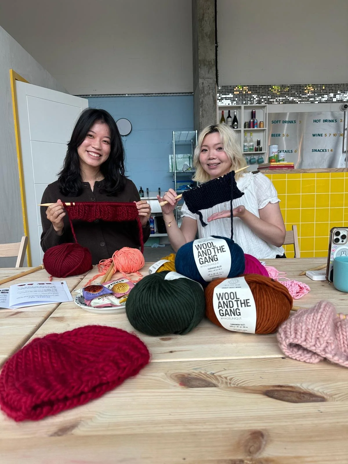 Two women learning to knit at a knitting workshop for beginners in London with knitting supplies and yarn, smiling at the camera, in a cozy indoor setting.