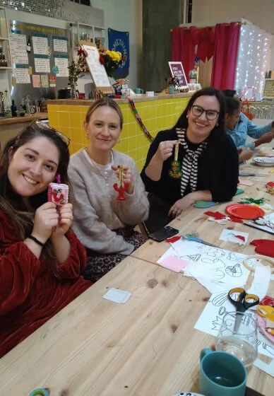 Three women in a craft workshop sitting at a crafting table smiling, holding handmade Christmas ornaments, with crafting supplies and festive decorations in the background.