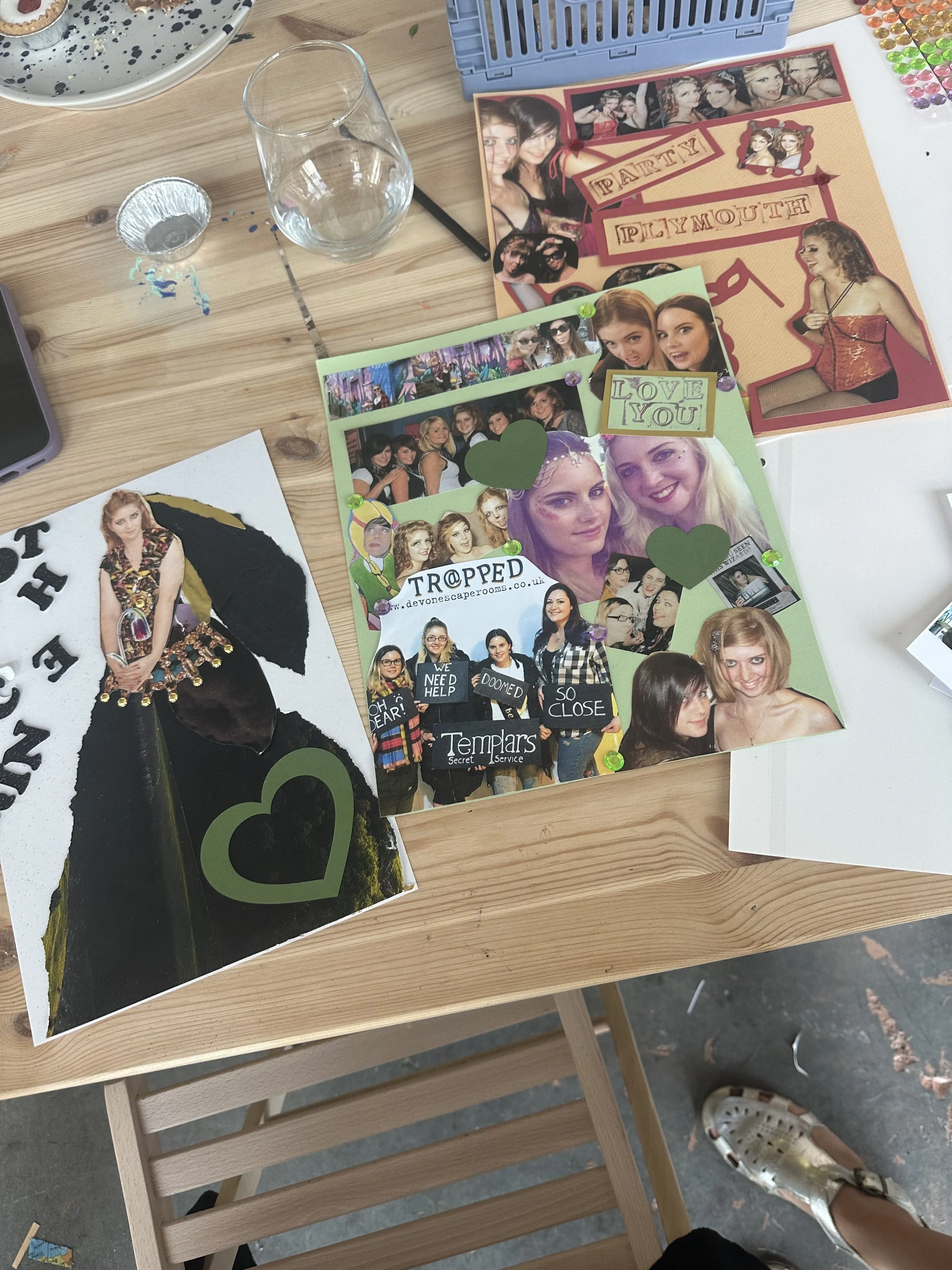 Table with various photo collages, a glass and a foil cup, and craft supplies. The collage in the front features people at a party with words like 'TROPPED,' 'LOVE YOU,' and signs saying 'OH DEAR' and 'WE NEED HELP.' The middle collage shows dancing