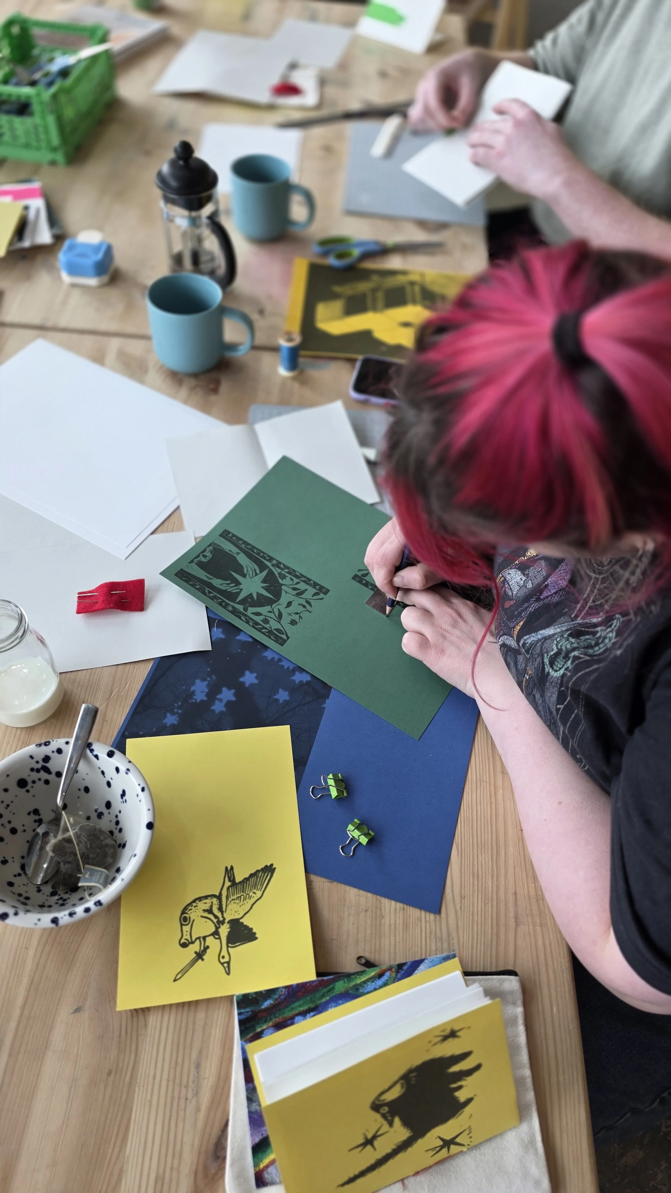 A person with pink hair at a bookbinding workshop in London is working on a craft project at a wooden table, surrounded by colourful papers, books, and stationery tools, including scissors, binder clips, and glue.