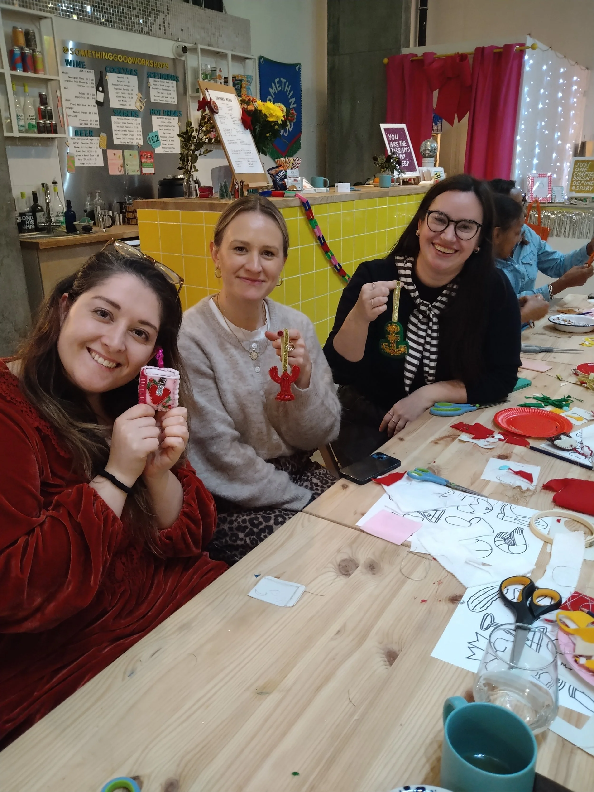 Three women sitting at a wooden table, holding decorated Christmas ornaments they made during a craft activity. The table has craft supplies like scissors, fabric pieces, and paper templates. The background shows a yellow-tiled counter, a menu, and festive decorations.
