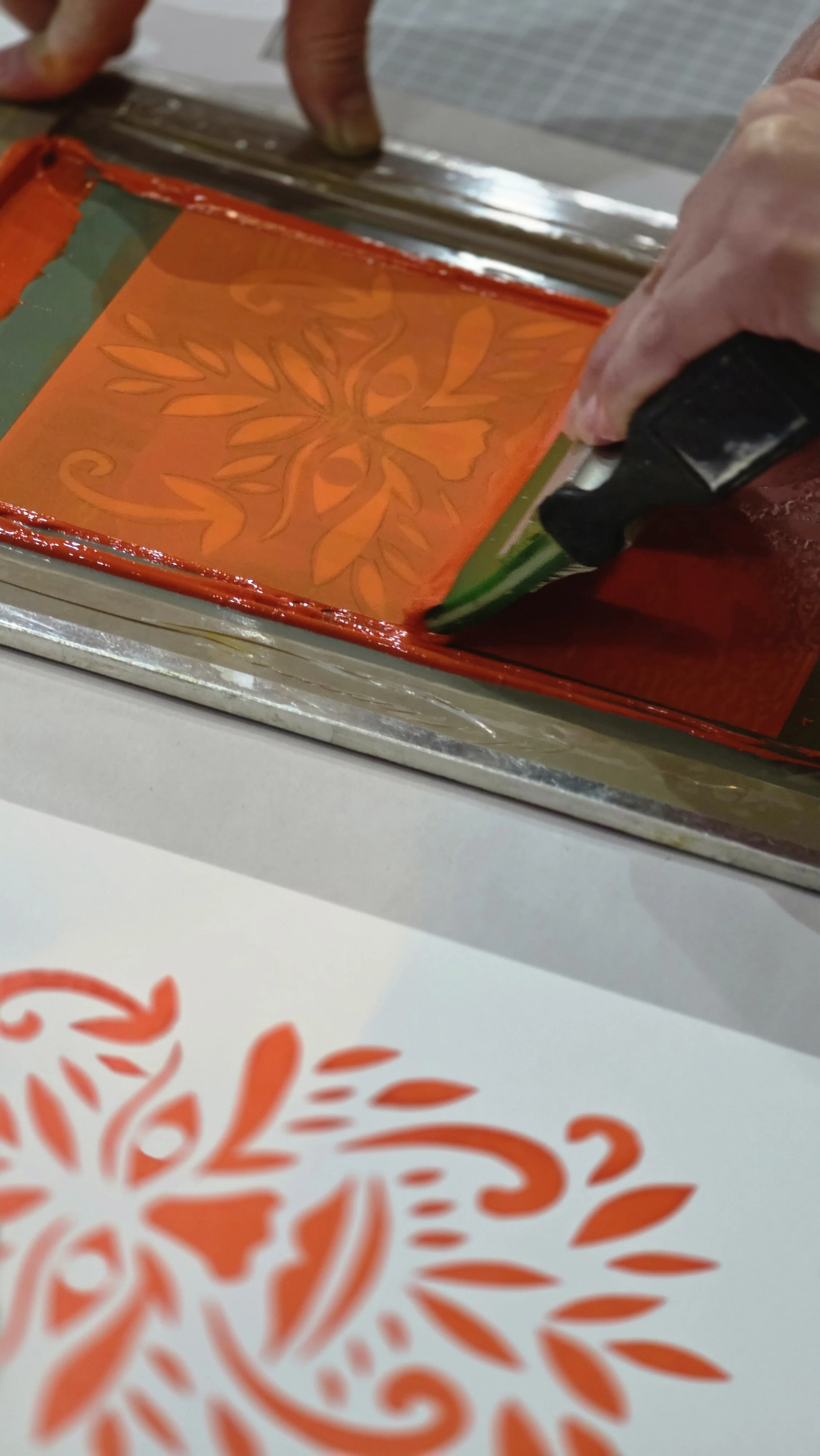 A person pulling an orange coloured screenprint of a floral design on a sheet of paper at a screenprinting workshop in London.