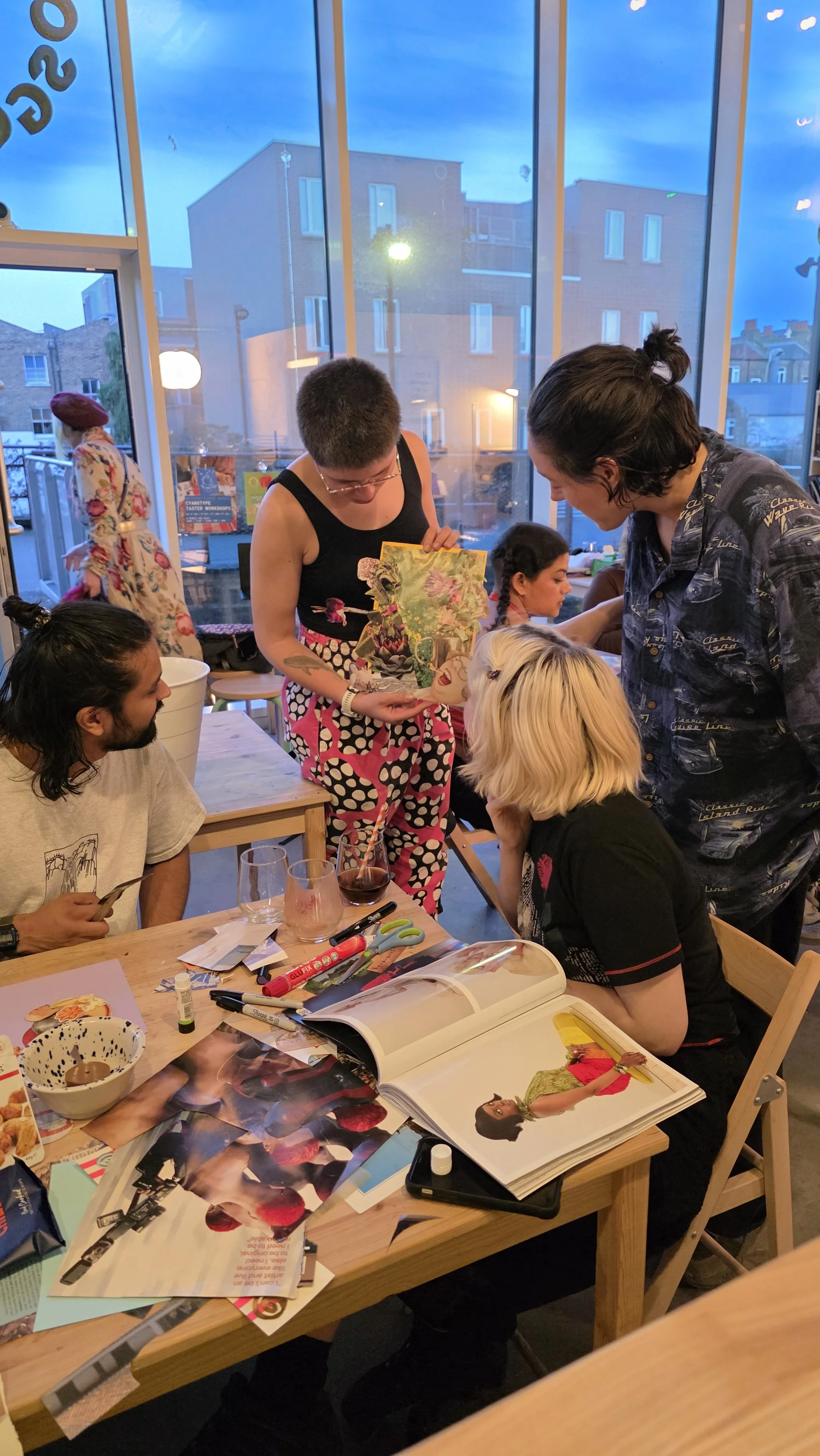Group of people gathered around a table with magazines and art supplies inside a craft workshop space at a collage night, with large windows showing a city street outside at dusk.