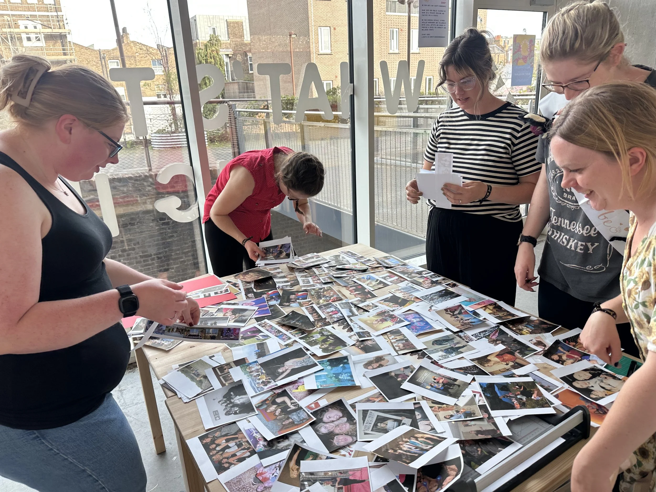 A group of five women looking at a table covered with printed photographs inside a glass-walled room, with buildings and a fence visible outside.