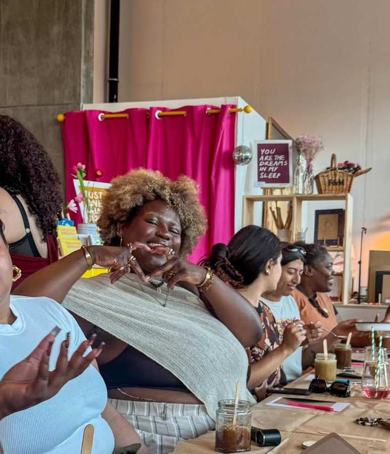 Group of diverse women sitting at a table, enjoying drinks and socializing, in a cozy indoor setting with pink curtains, decorative signs, and various decor items at Something Good Workshops in Deptford, London.