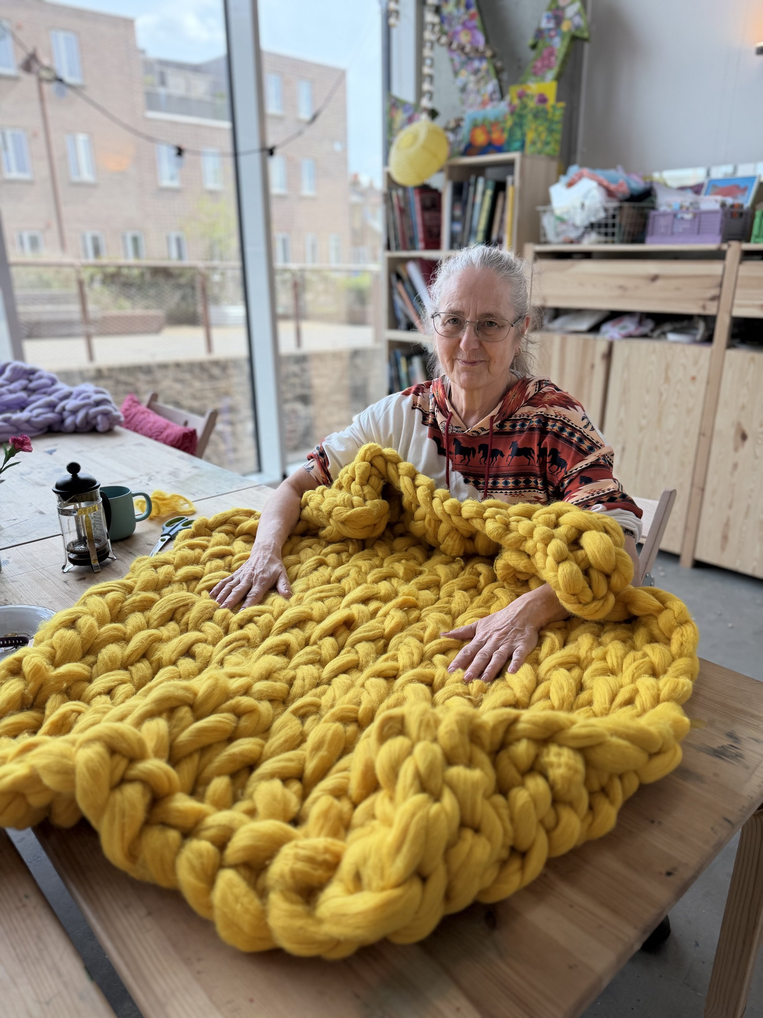 An elderly woman with glasses sitting at a table, touching a large, chunky yellow knitted blanket or yarn project at an arm knitting workshop in London.