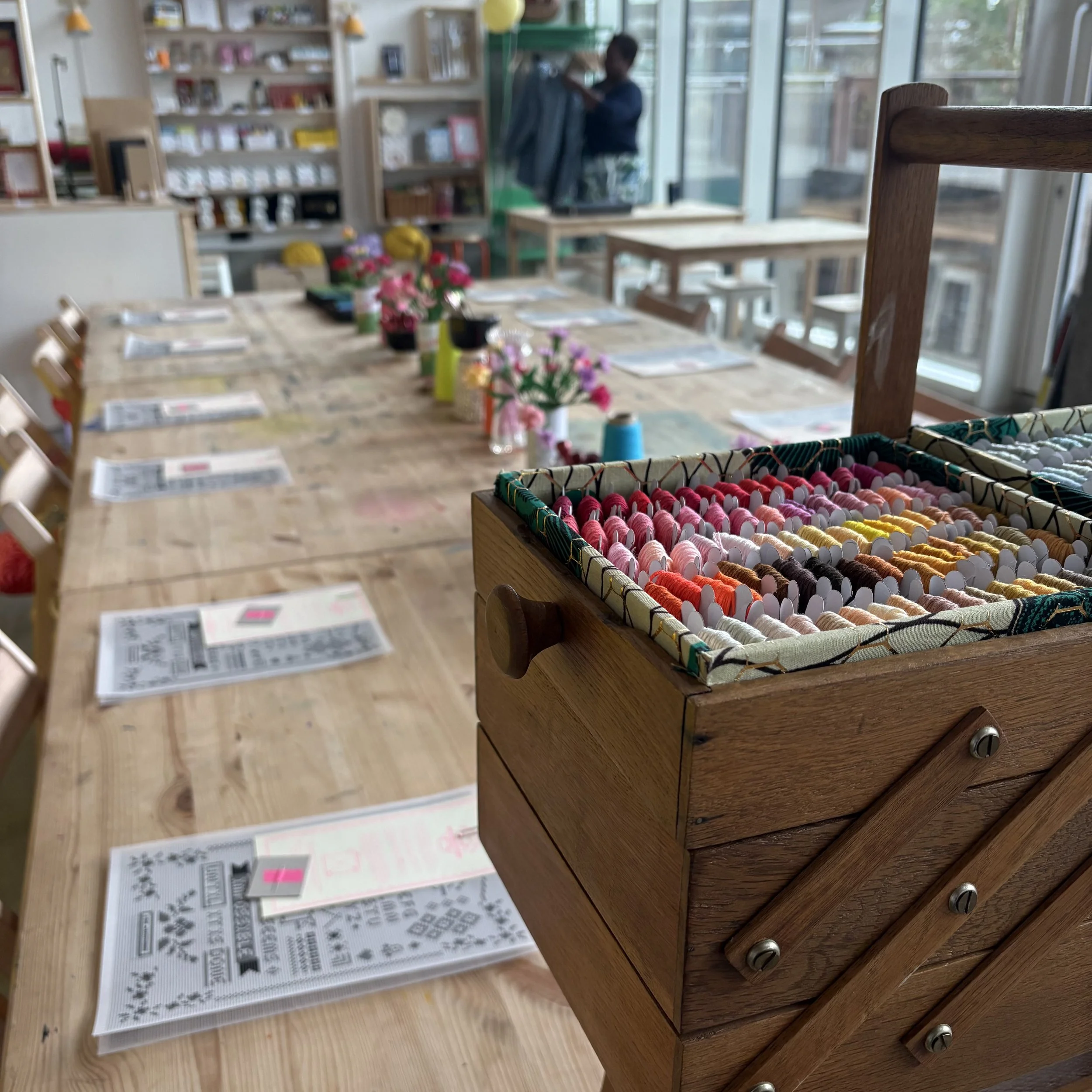 A wooden table with a sewing box containing a rainbow of embroidery threads, with printed knitting patterns and instructions nearby. In the background, a woman is standing near windows with shelves containing books and other items in a well-lit room.