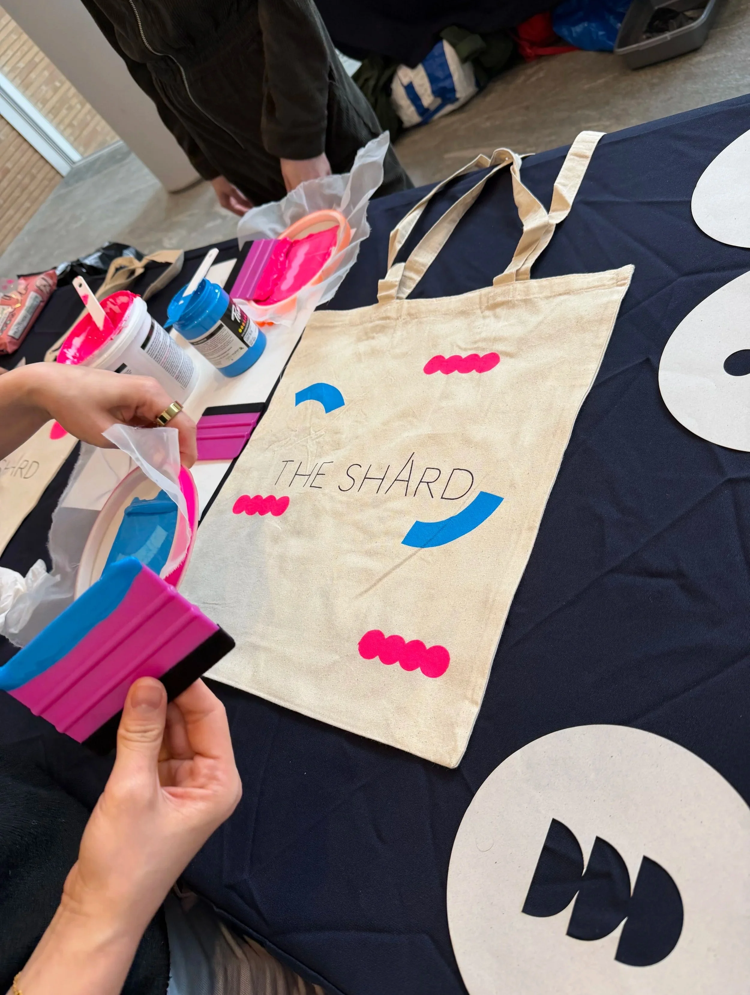 Person holding pink and black paper cutouts on a table with various items including a beige tote bag with the words "The Shard" written on it.