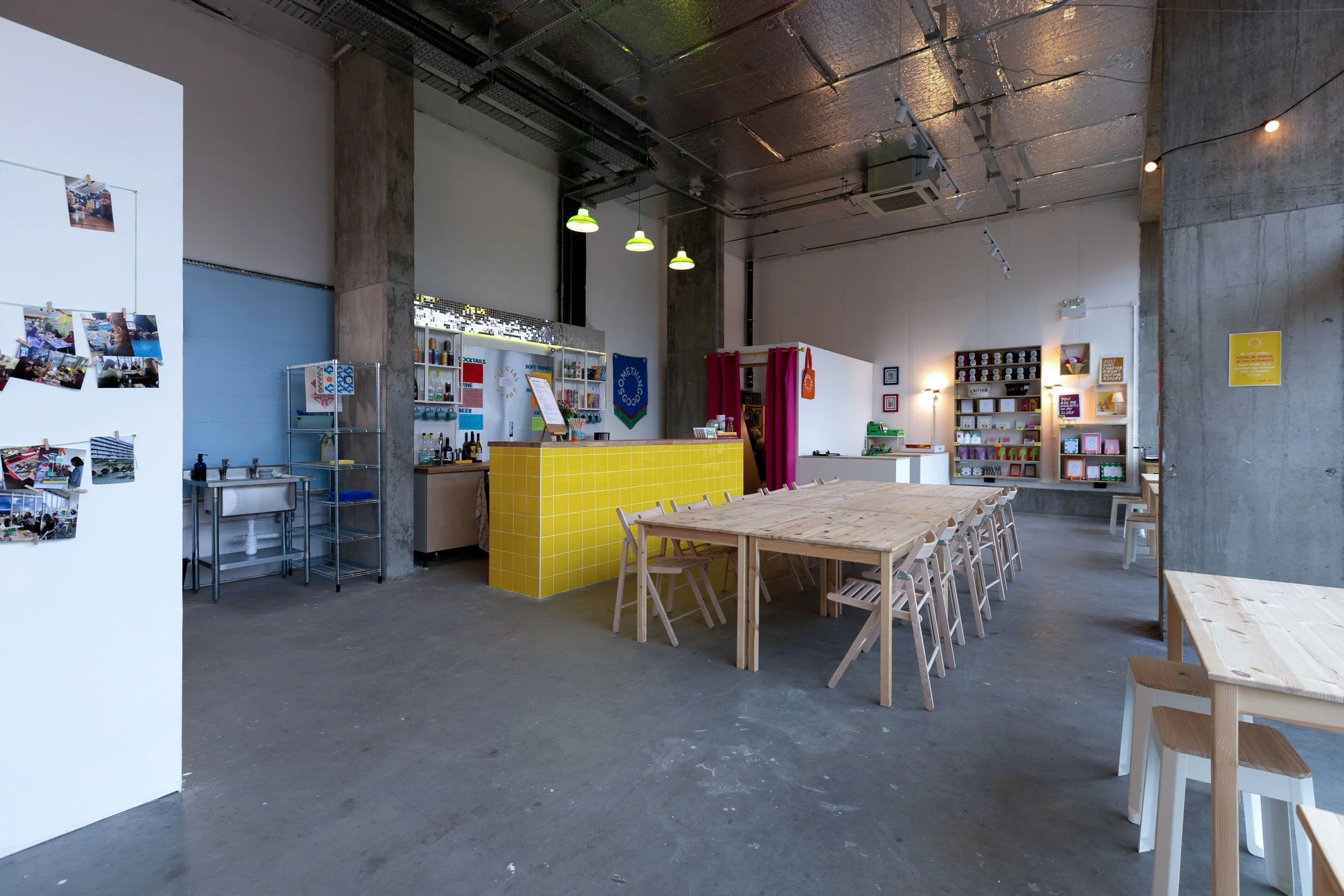 Interior of Something Good Workshops in Deptford, London with a long wooden table and chairs, a yellow-detailed counter area, and shelves with books and decor. The ceiling has exposed industrial elements with green hanging lights.