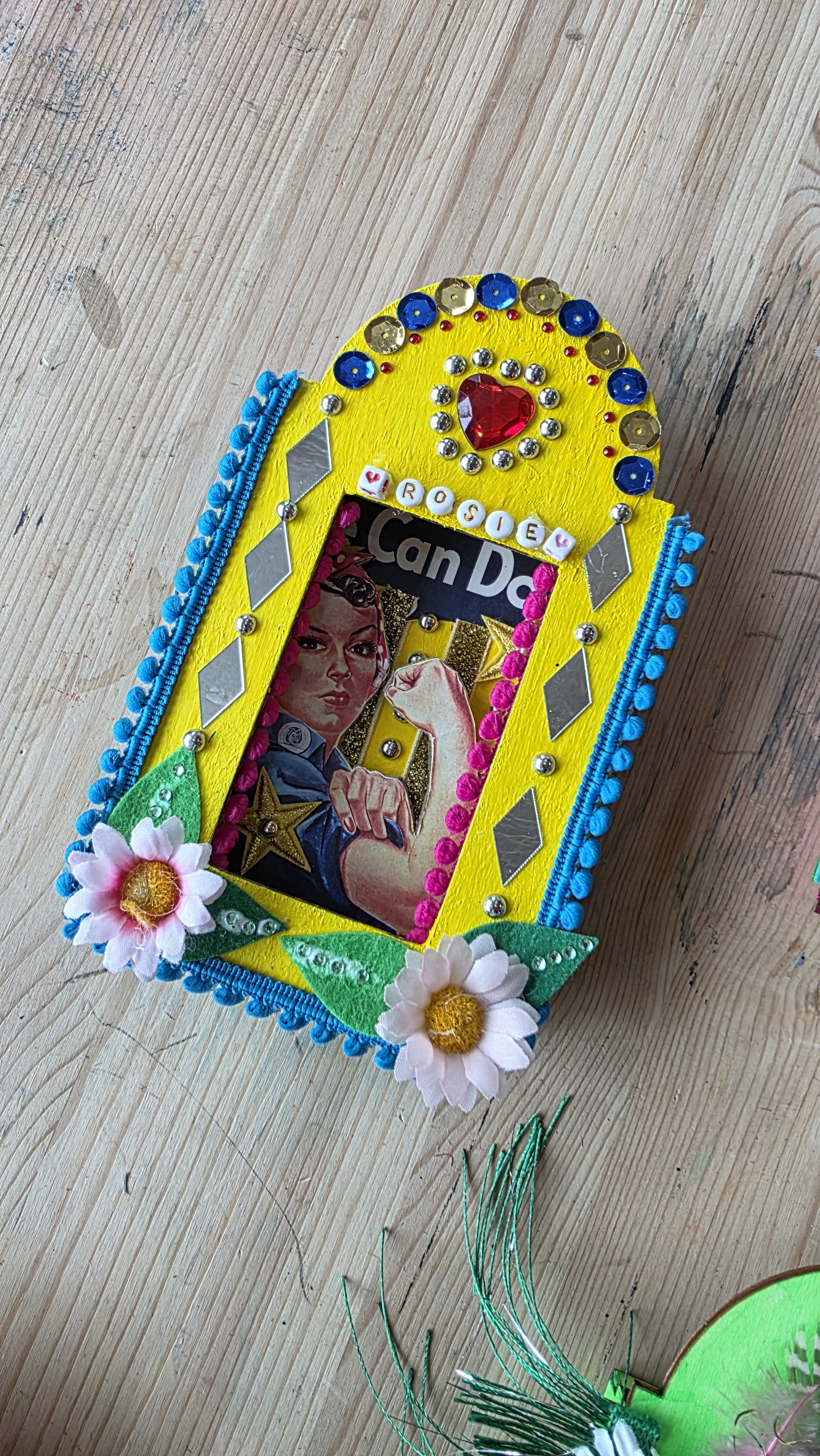 Decorative handmade craft shrine with flowers, beads, sequins, and magazine cutouts, on a wooden surface.