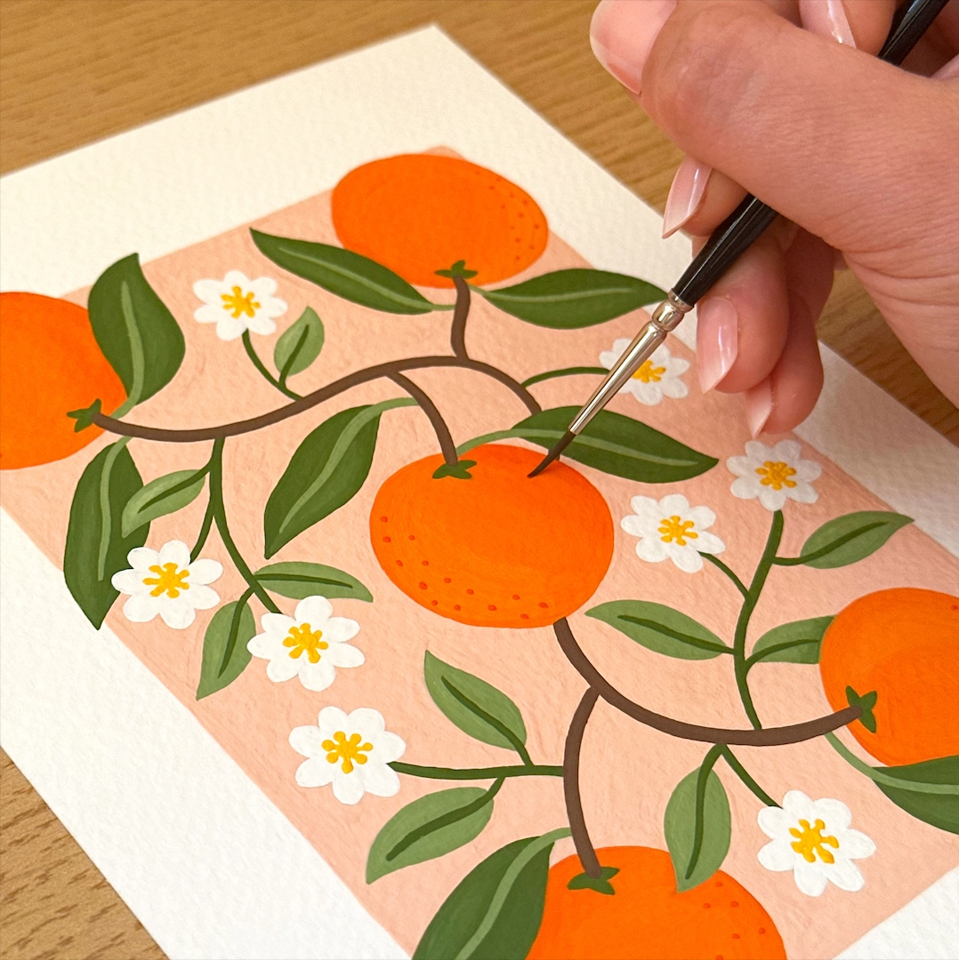 Person painting a pattern of oranges, green leaves, and white flowers on paper with a fine brush.