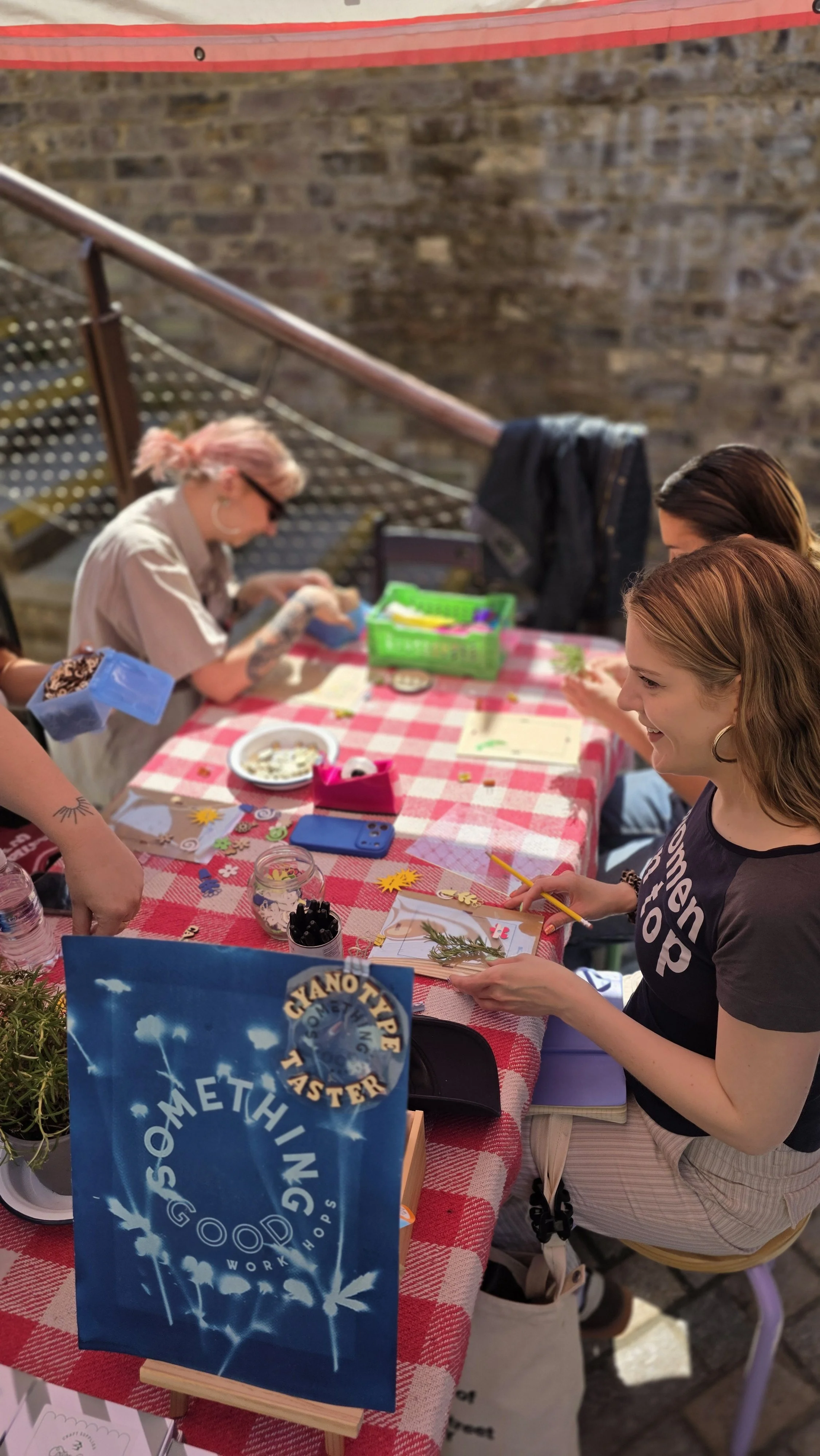 People participating in a crafting workshop at a table with a red and white checkered tablecloth, various craft supplies, and a blue sign that says 'Cyanotype Taste Workshop'. The setting is an outdoor market near a brick wall.