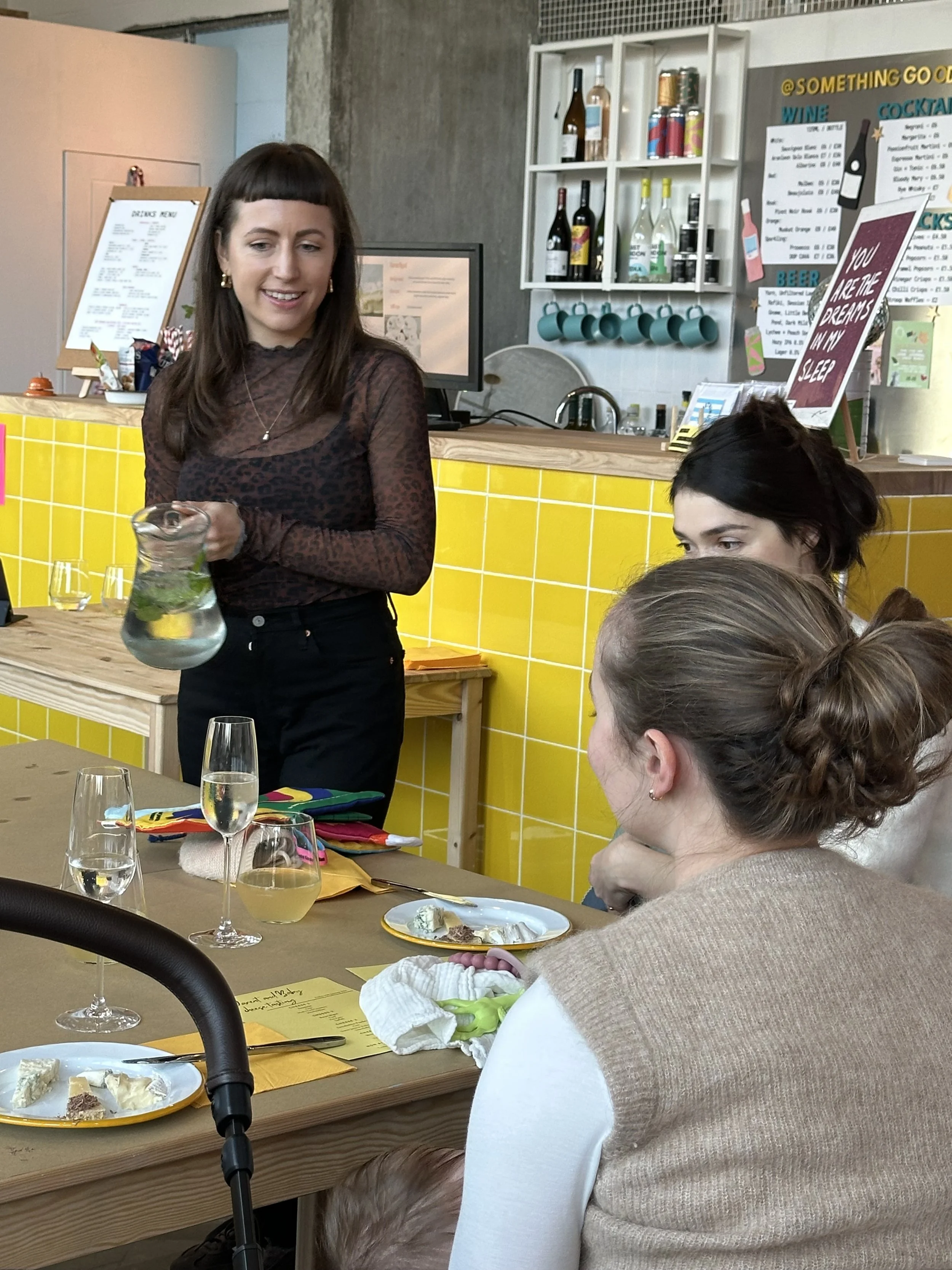 A woman standing and smiling, holding a clear glass pitcher with lemons and mint leaves at a table with glasses, plates, and a yellow paper, while three women sit at the table in a restaurant with yellow tile walls and a menu in the background.