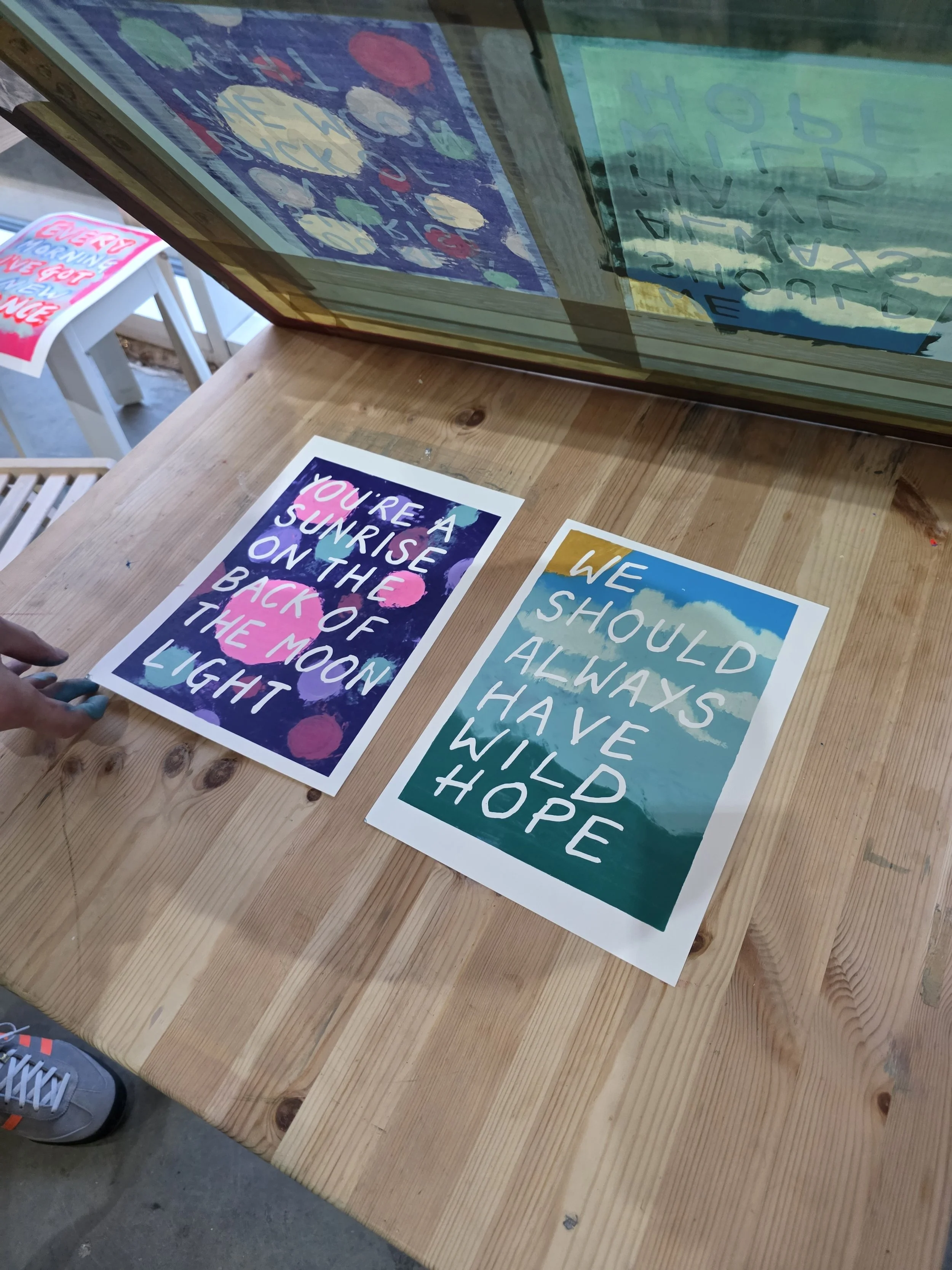 Colorful Adam Bridgland monoprint screenprint posters with inspirational messages on a wooden table, with reflections visible on the surface of a glass case above.
