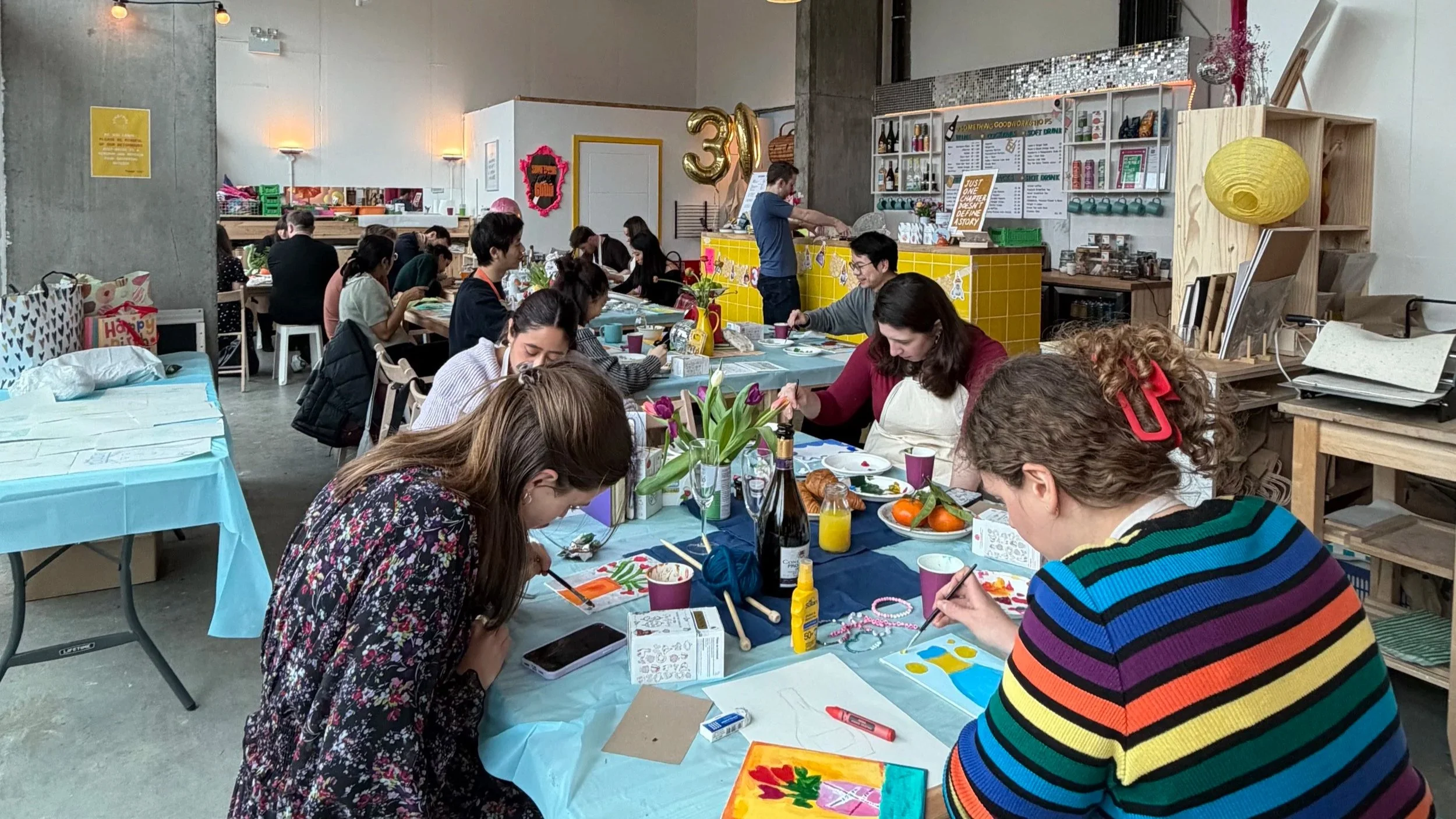 Two women standing together inside a room, smiling, with one wearing an orange chunky knit shawl and the other a teal chunky knit shawl. There are books, pink tissue paper, and green and purple baskets on a wooden shelf behind them. On the wall, a blue cloth with white text reads 'SOMETHING GOOD,' and a large decorative flower with white petals and a yellow center is mounted on a plywood wall.