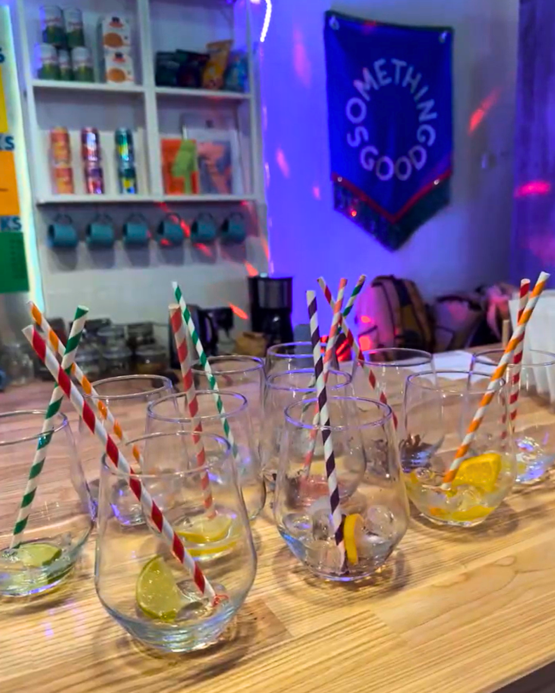Several glasses with lemon slices and striped straws on a wooden counter, with a bar or kitchen area and a 'Something Good' banner in the background.