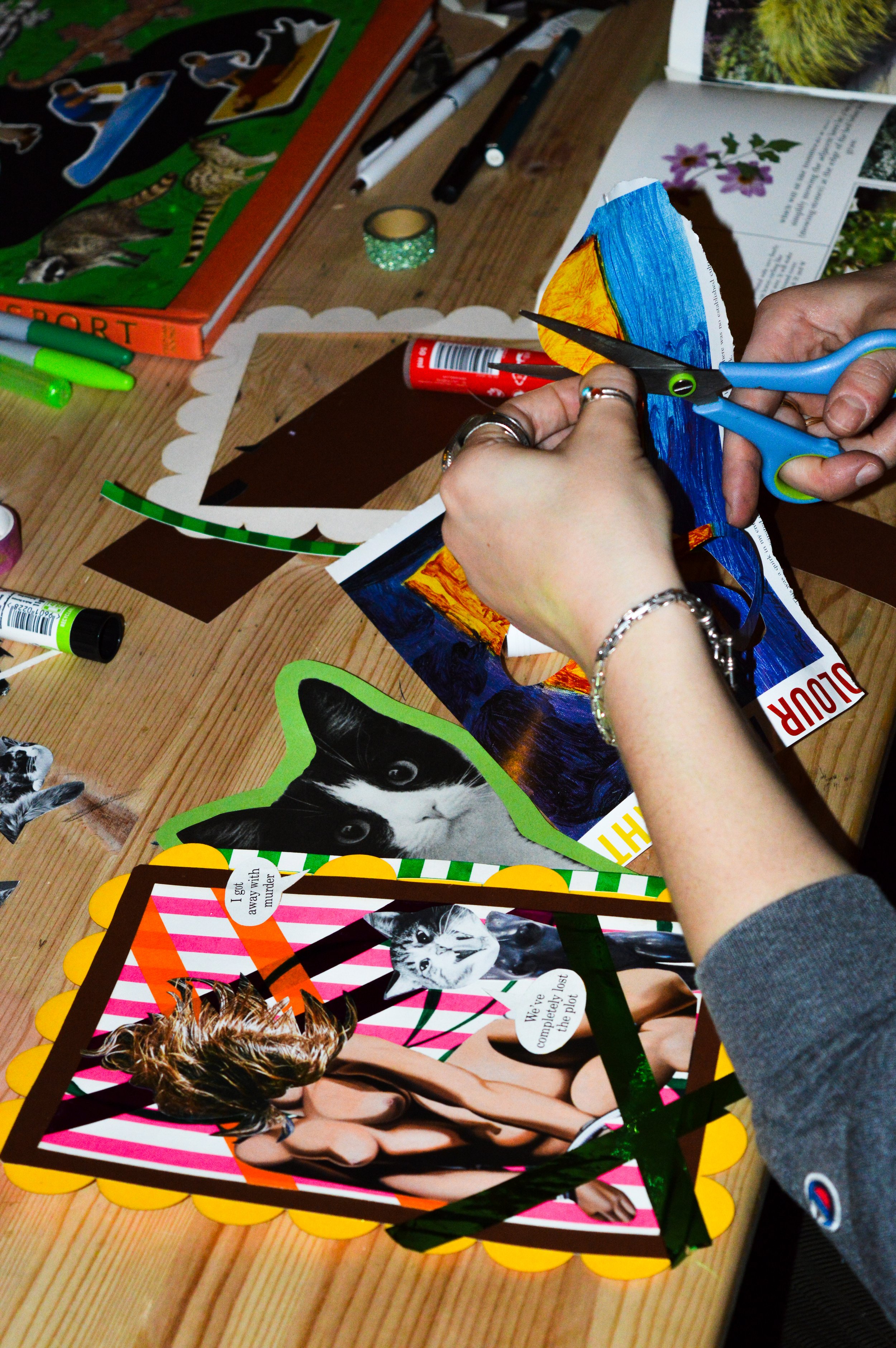 Person cutting out a colorful, collage from a book at a cluttered wooden table filled with markers, stickers, and other colorful papers.