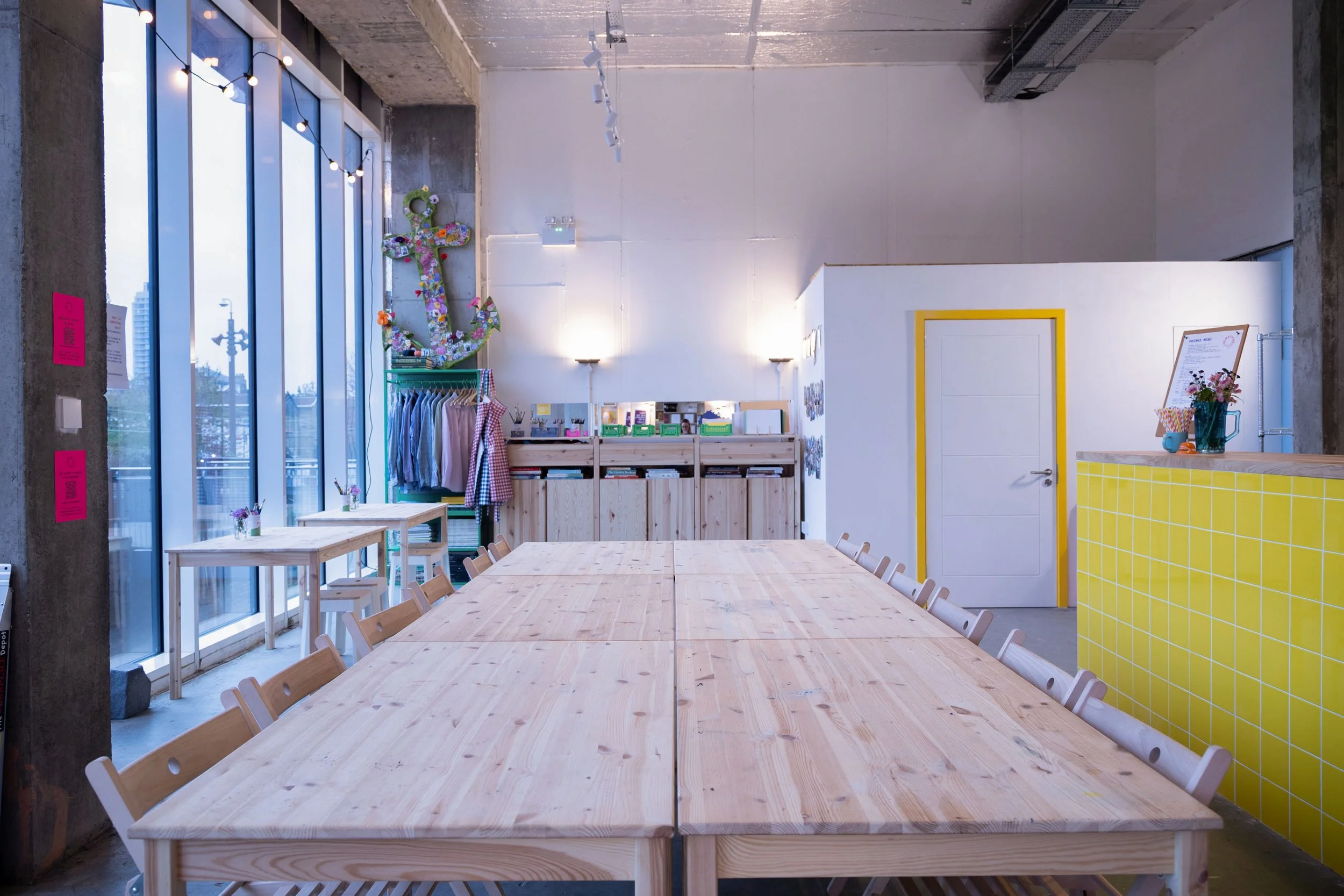 A modern, minimalist interior with a long wooden table and chairs, large window panels with string lights, and decorative elements including a colorful anchor sculpture and umbrellas, bright yellow counter, and bookshelves.