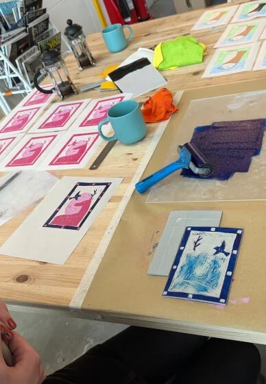A workspace with a wooden table covered in art supplies, paper, and images. There is a blue mug, a green cloth, an orange cloth, a soapstone, and a foam roller on the table. The images feature flamingos and birds in various colors, and a roller is being used to apply a purple ink or paint onto a piece of paper.