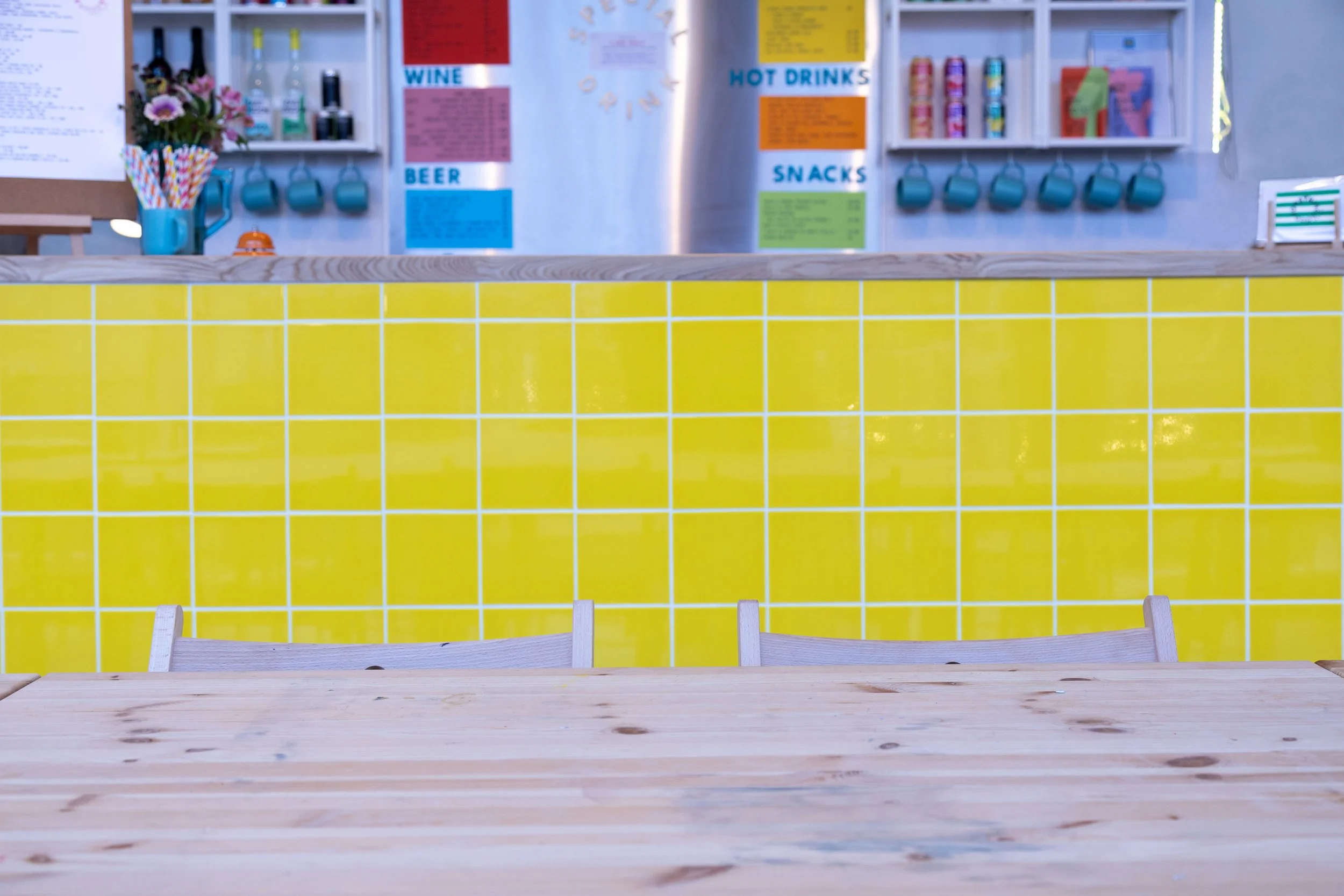 A counter with a yellow tile front and a wooden top, with shelves in the background displaying colorful signs, mugs, and products at a café or shop.