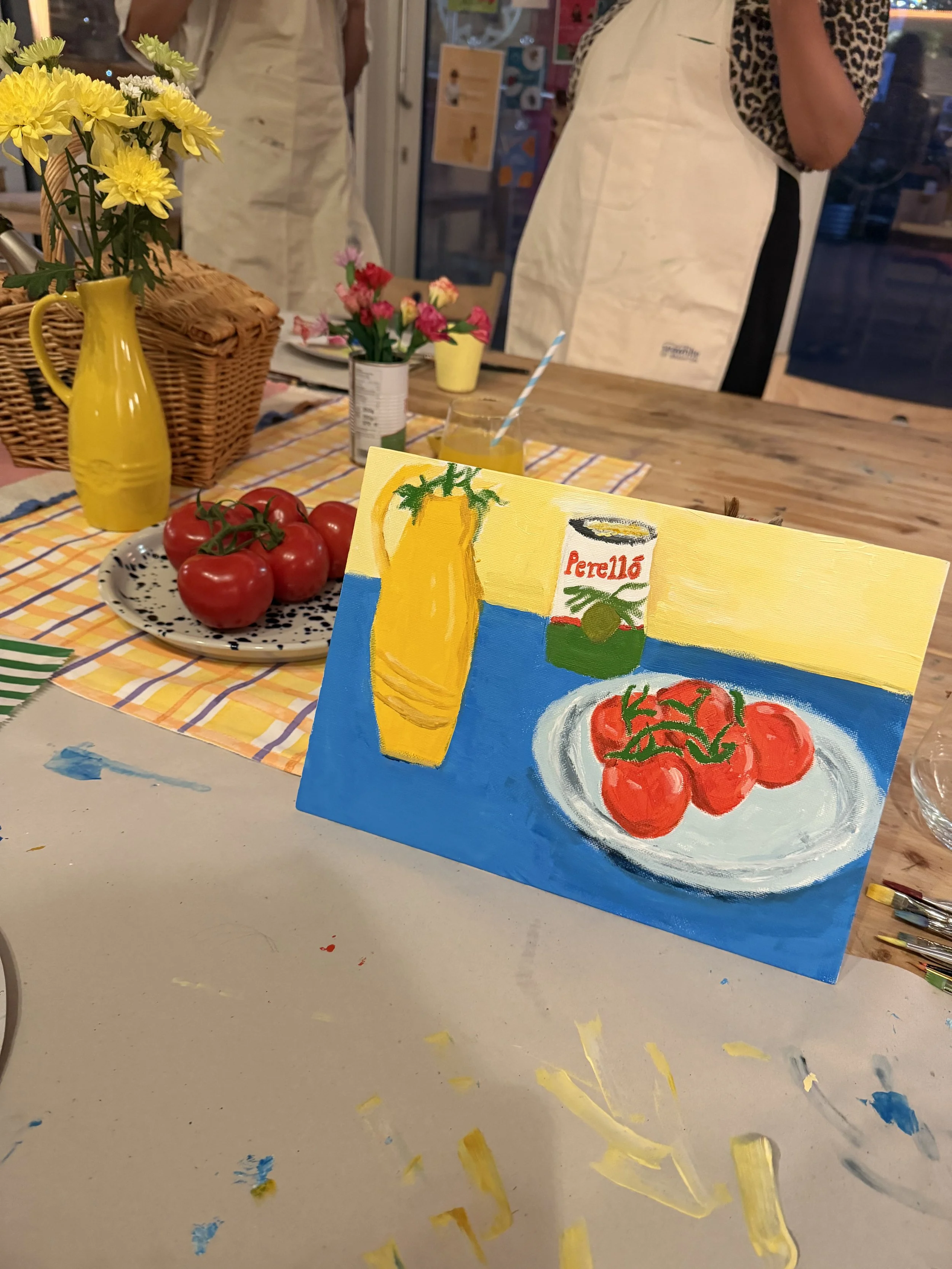 A colorful painting depicts a yellow pitcher and a plate of tomatoes on a table set for painting. In the background, people are gathered around a table with flowers, plates, and drinks, in a cozy indoor setting at a still life painting workshop.