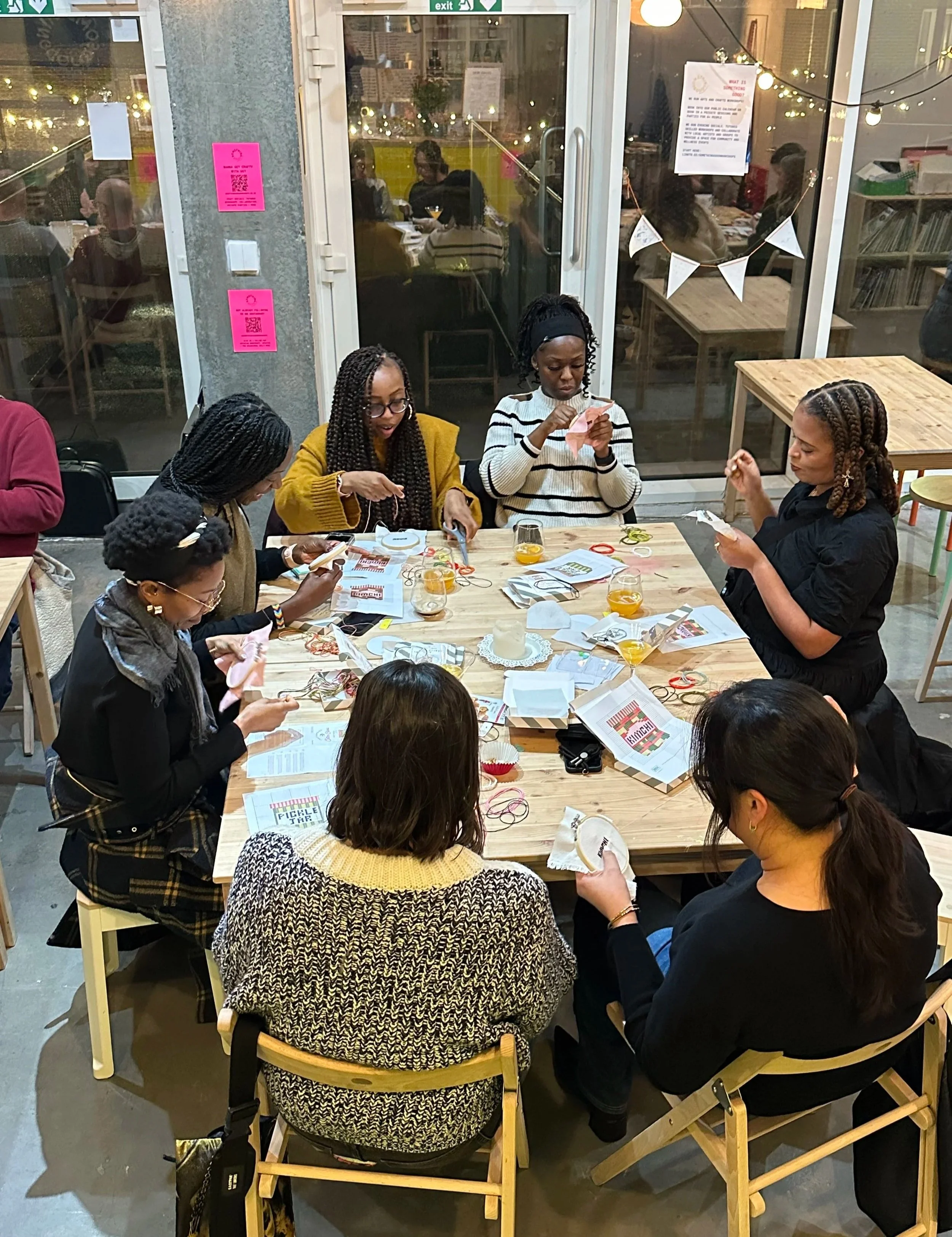 A group of women are sitting around a wooden table engaged in arts and crafts activities. The table has various craft materials, papers, and glasses of orange juice. Through the large glass window behind them, several people can be seen sitting inside a brightly lit room, and the reflections of string lights and some signs are visible on the glass.