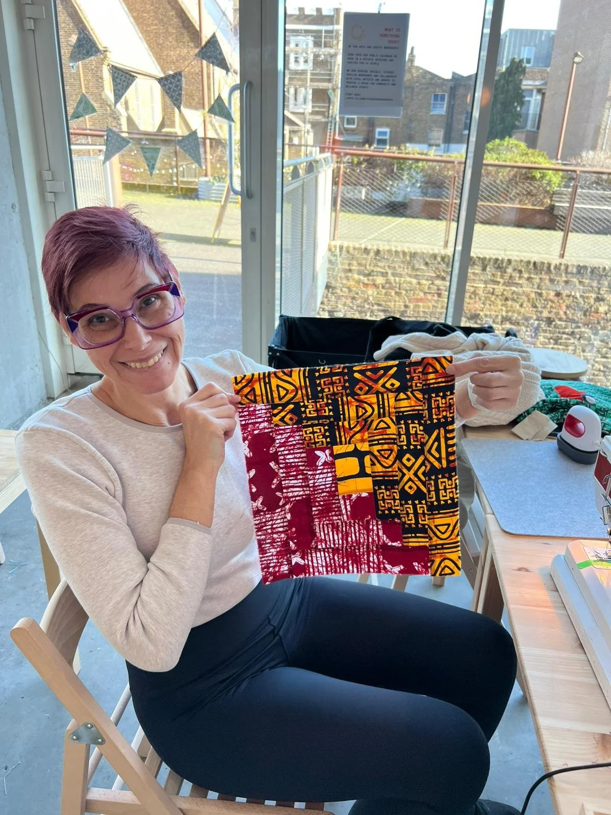 A woman with short purple hair, glasses, and a white shirt is sitting on a chair, smiling and holding up a piece of fabric with colorful patterns. She is indoors next to a window, with various sewing supplies and fabrics on a table nearby.