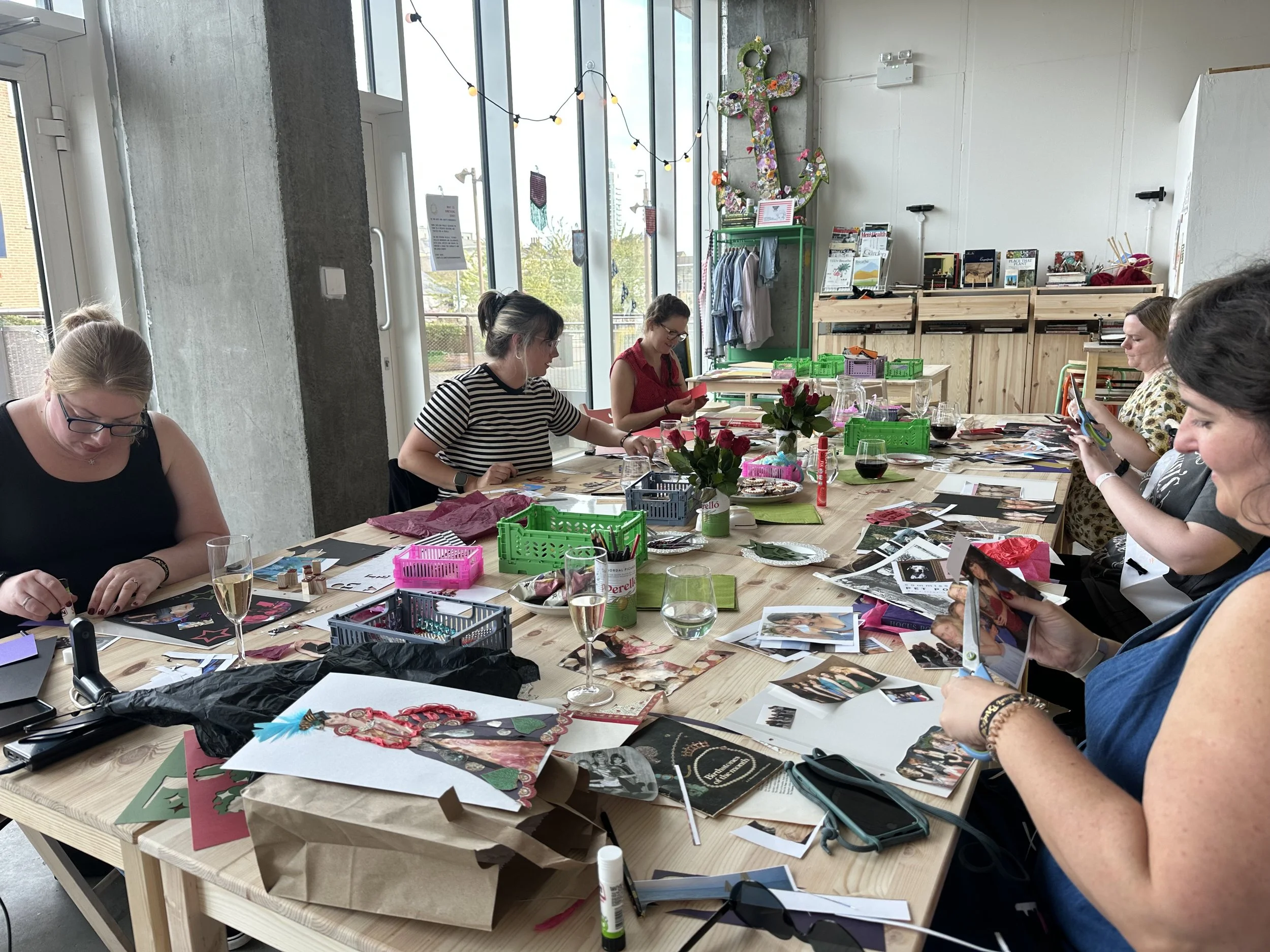 Women sitting at a long table working on arts and crafts projects with various materials, photos, and decorations, in a bright room with large windows and plants.