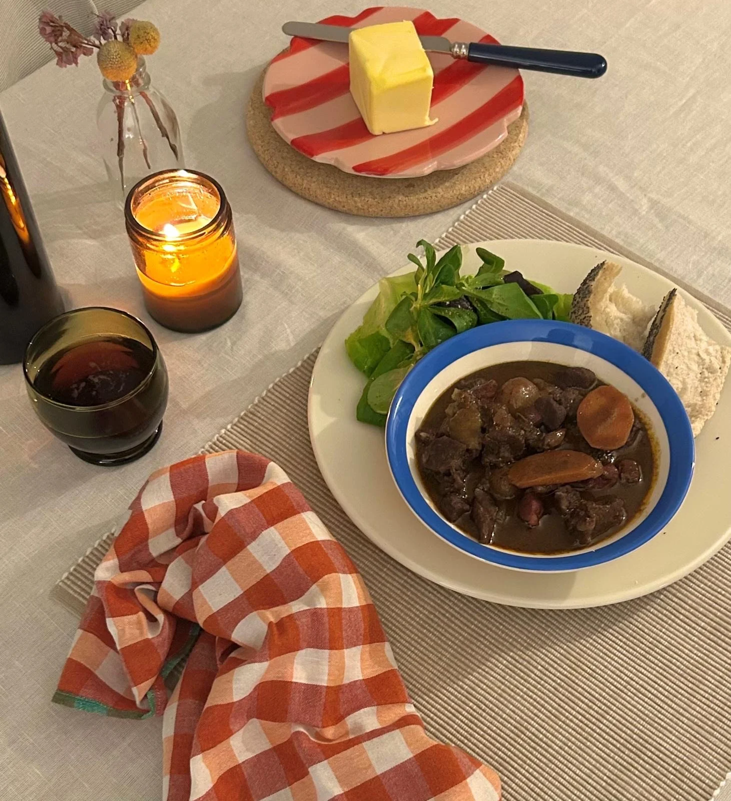 A dinner table with a bowl of beef stew, a salad, and bread. There is a lit candle and a glass of red wine on the table. A small flower vase with dried flowers is also present. A plate with butter and a knife is in the background.