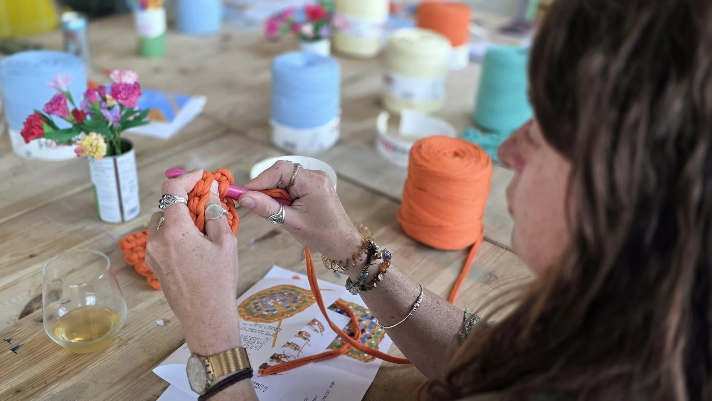 A woman with bracelets and rings is learning to crochet with orange yarn at a crochet workshop for beginners in London, with colorful yarn rolls and flowers on the table.