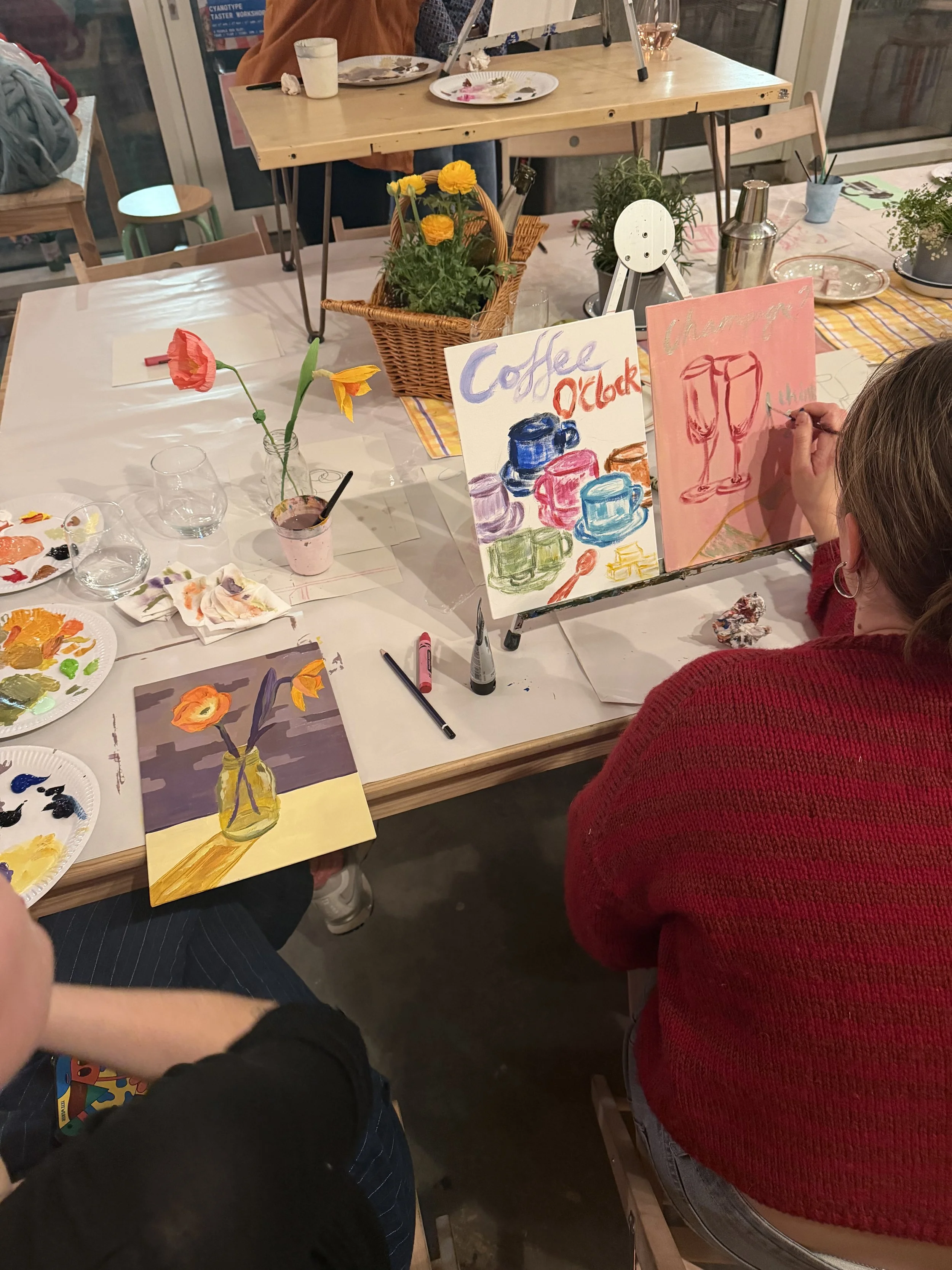 People creating colorful paintings at a craft table with brushes, paint, and drawings of flowers and glasses.