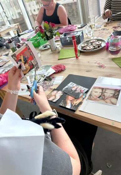 People sitting at a table decorating photo albums with scissors, glue, and printed photos, with flowers and drinks on the table.