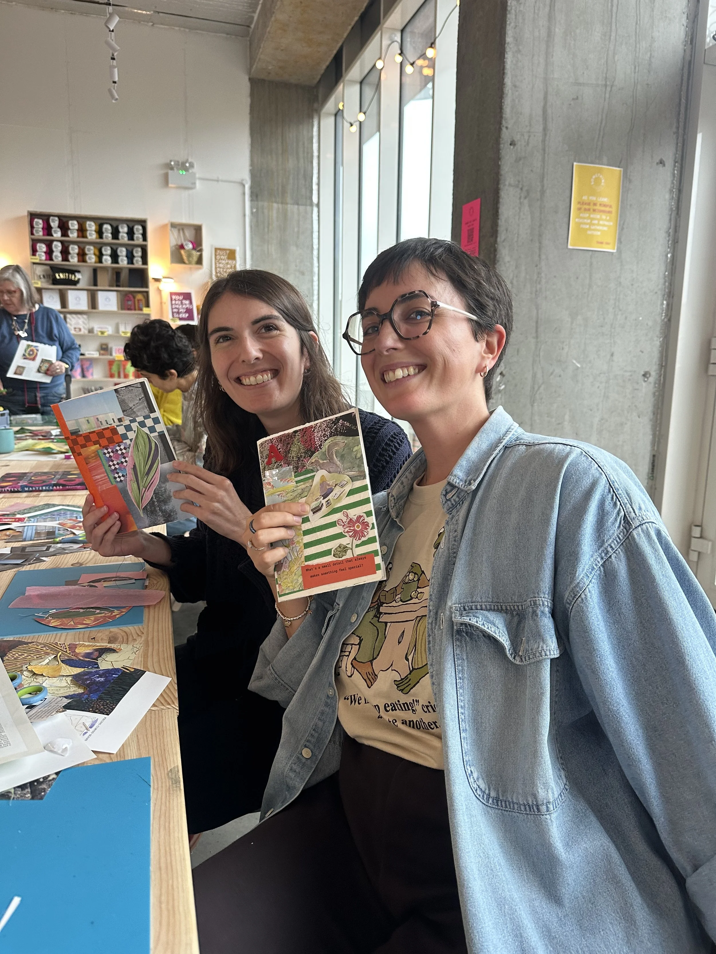 Two women smiling and holding colorful junk journals craft workshop in London. There are other people, craft supplies, and artwork in the background.