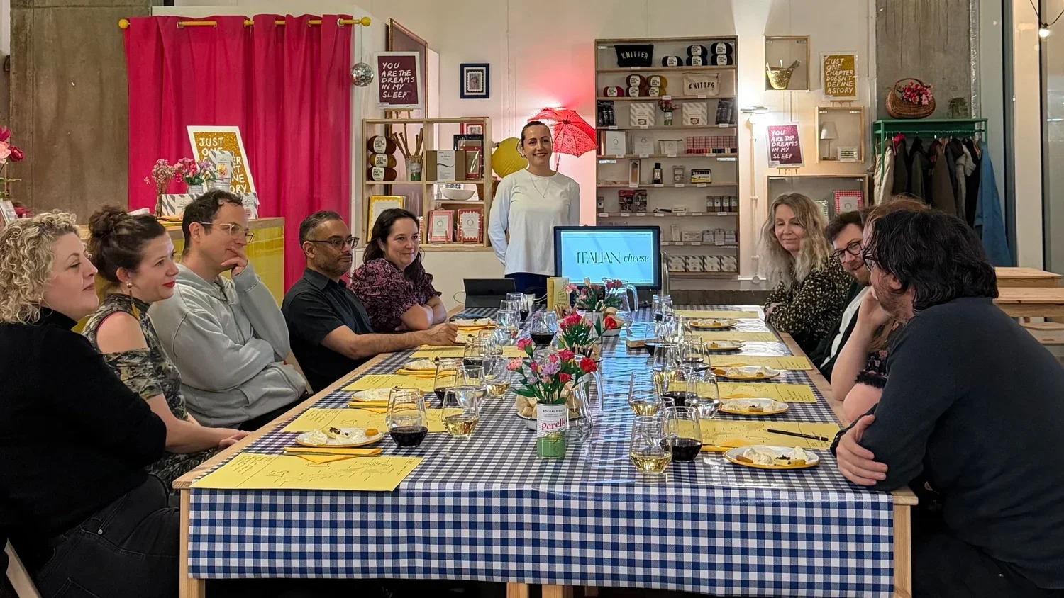 A group of people seated around a long table with food and drinks, attending a cheese tasting event in a cozy, decorated room.